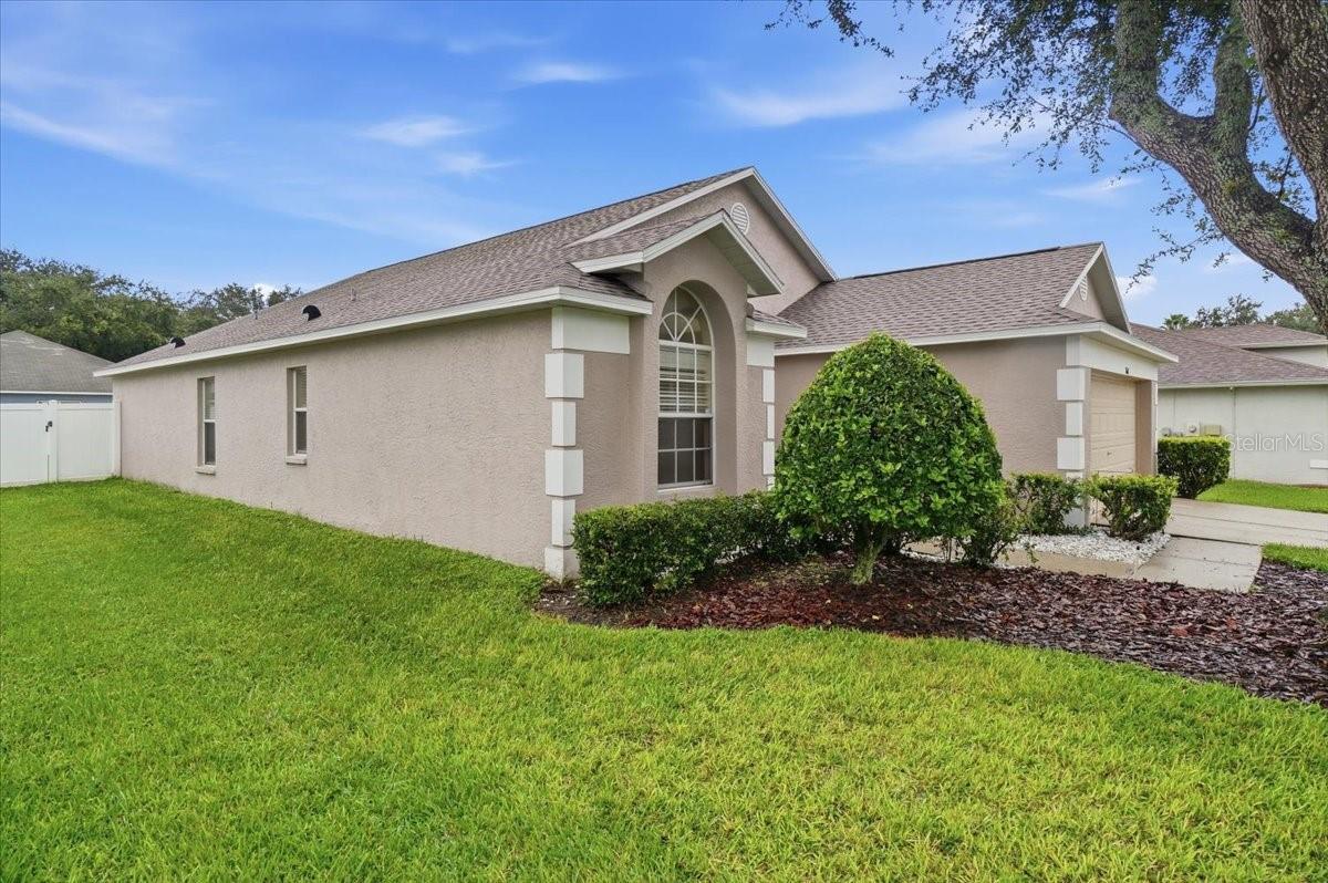 Single-story beige stucco home with an arched front window, trimmed shrubs, and a broad green lawn.