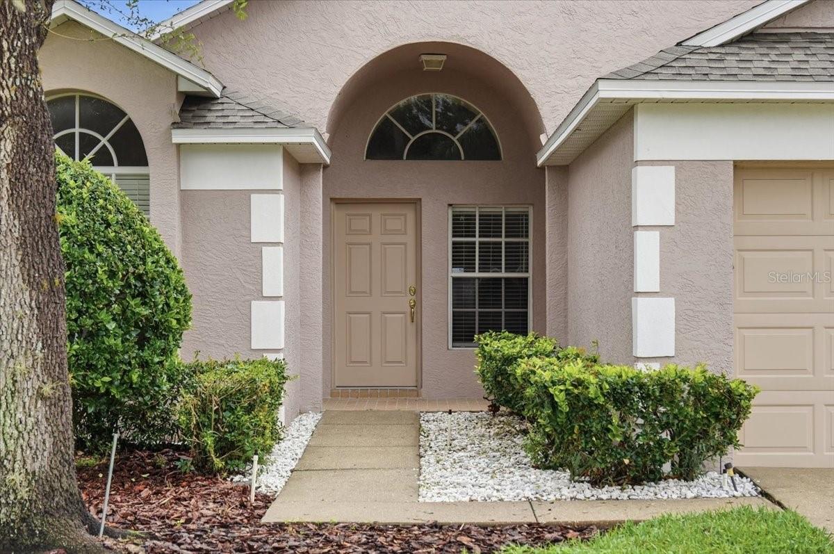 Stucco home entrance with arched windows, a tan front door, and landscaped walkway.