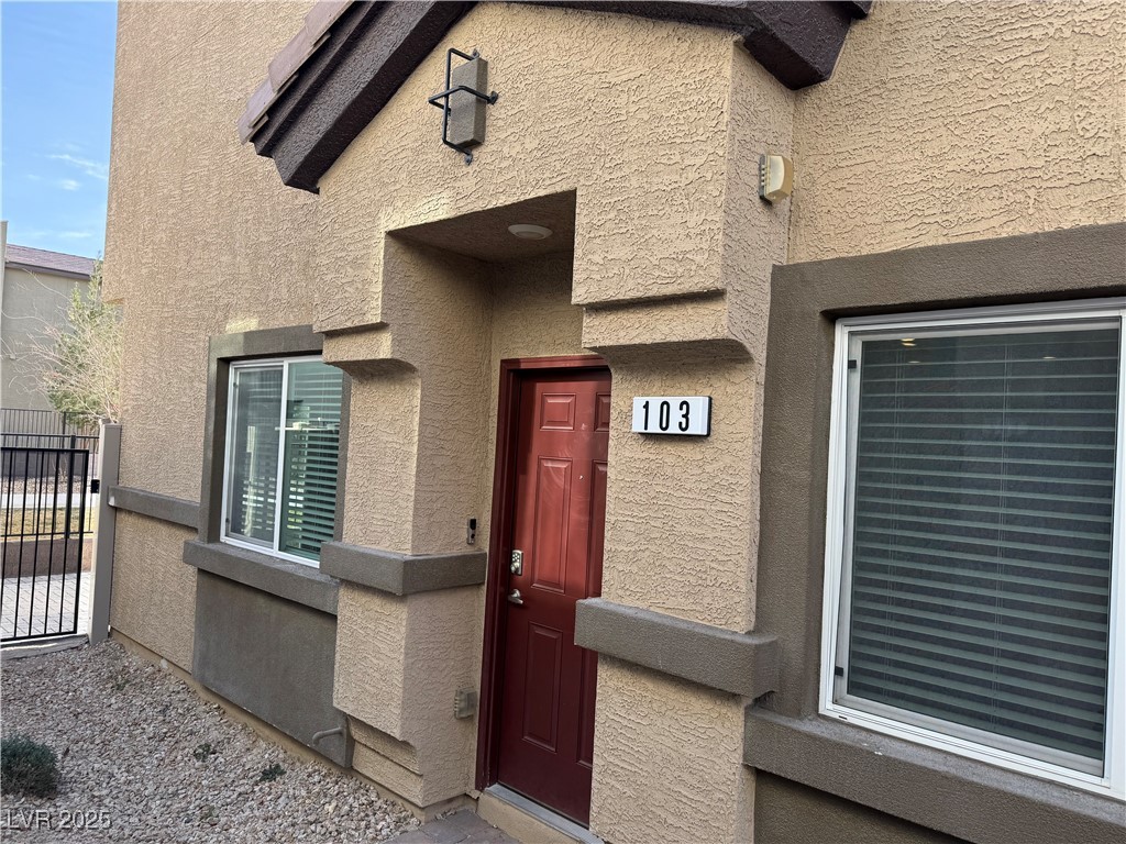 Tan stucco home entrance with a red door marked 103 and windows with blinds.