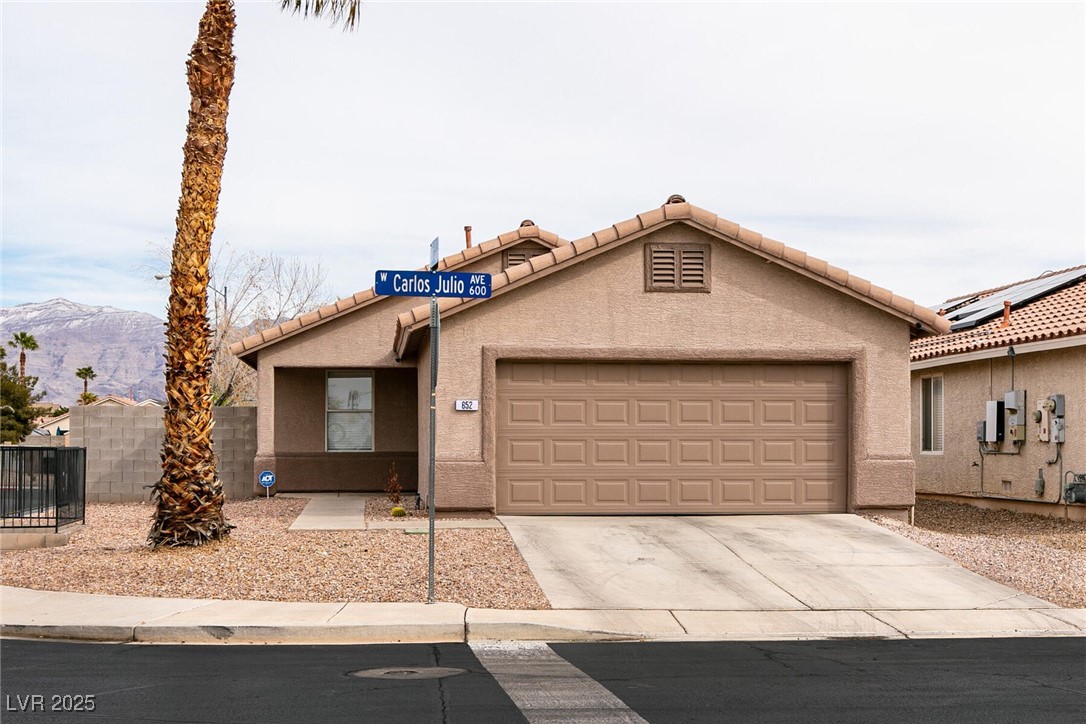 Single-story tan house with a two-car garage, gravel yard, palm tree, and distant mountains.