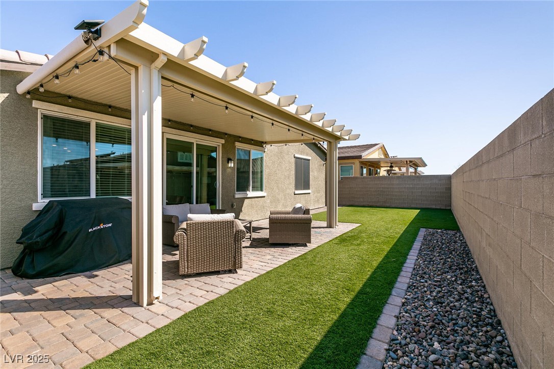 Fenced backyard with a pergola-covered paver patio, wicker seating, and a narrow green lawn.