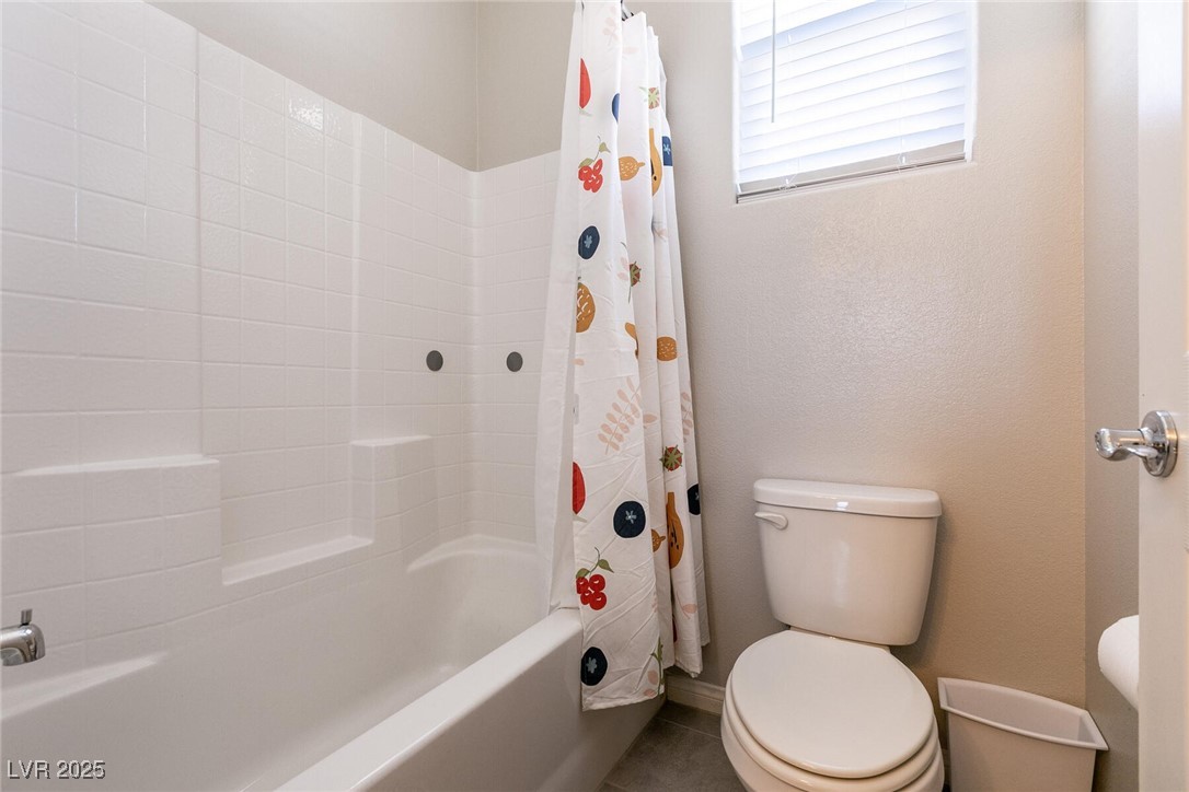 Bathroom with a white tiled tub and shower, patterned curtain, and toilet beneath a window.