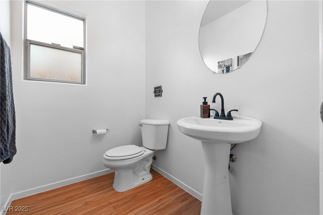 Bathroom with a pedestal sink, round mirror, frosted window, and wood-look flooring.