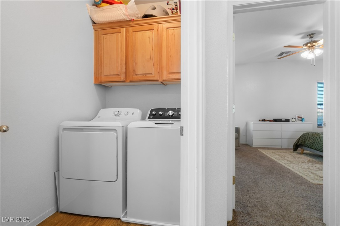 Laundry room with washer and dryer beneath wooden cabinets, opening to a carpeted bedroom.