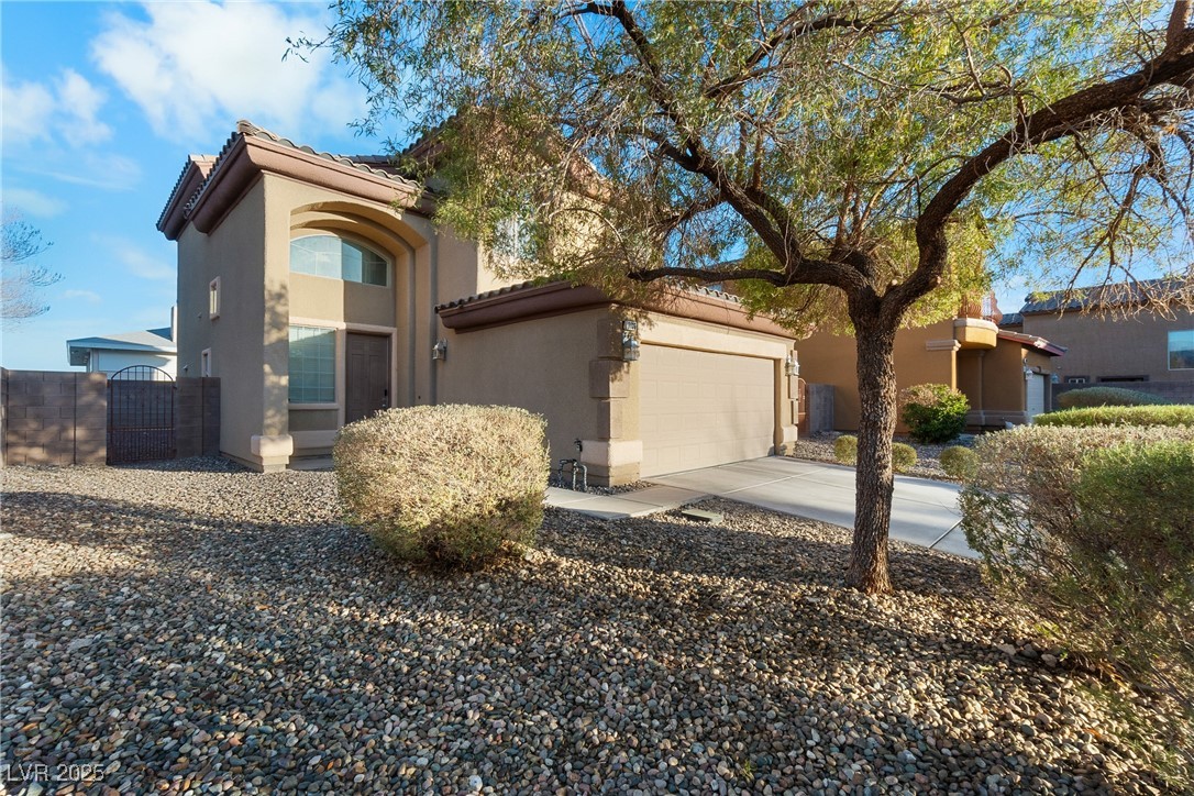 Two-story tan stucco home with an attached garage, gravel landscaping, and a large shade tree.