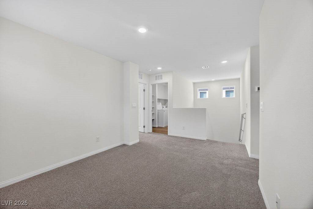 Empty carpeted room with recessed lighting and a laundry area visible through an open doorway.