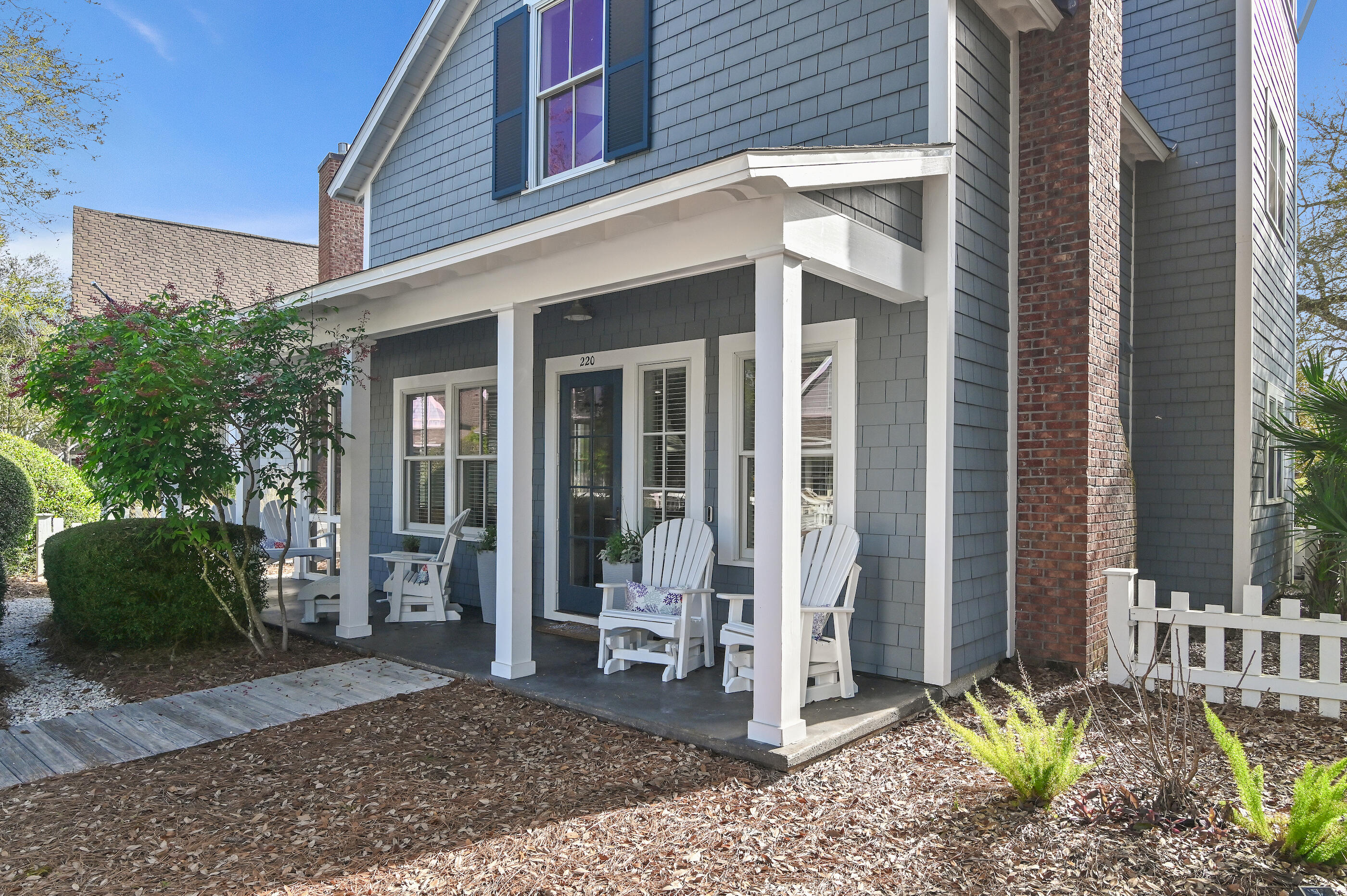 Blue-gray two-story home with a covered porch, white Adirondack chairs, and a prominent brick chimney.