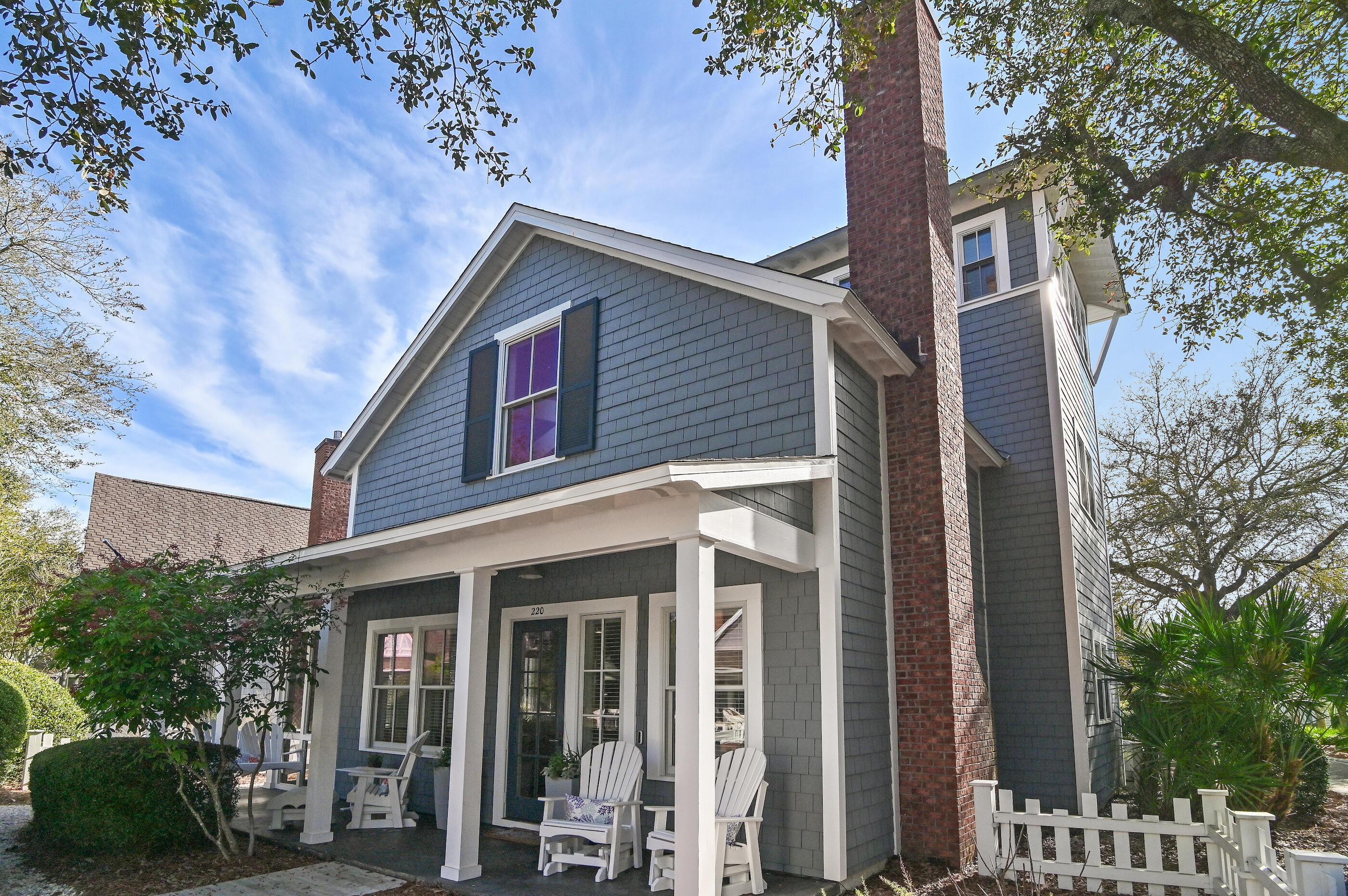 Gray-shingled two-story house with a covered porch, white Adirondack chairs, and a tall brick chimney.