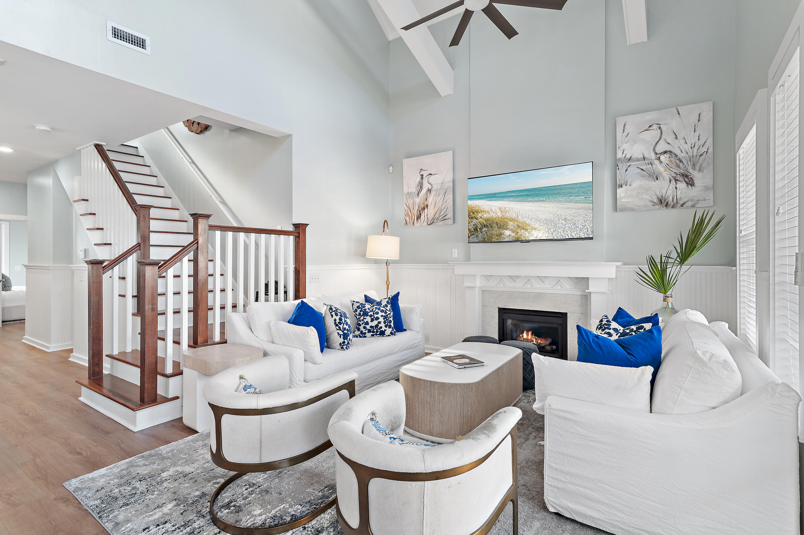 Living room with white sofas, blue pillows, a fireplace, and a dark wood staircase.
