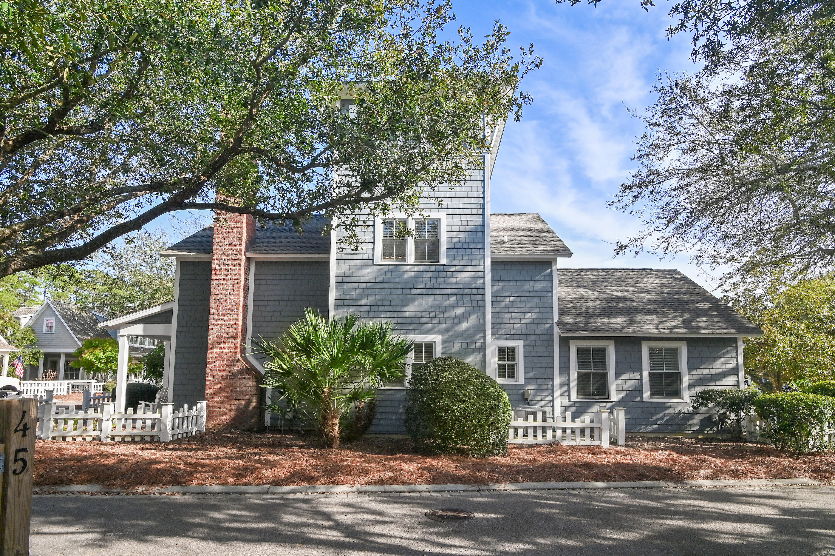 Blue-gray shingle house with white trim, red brick chimney, and white fencing beneath mature trees.
