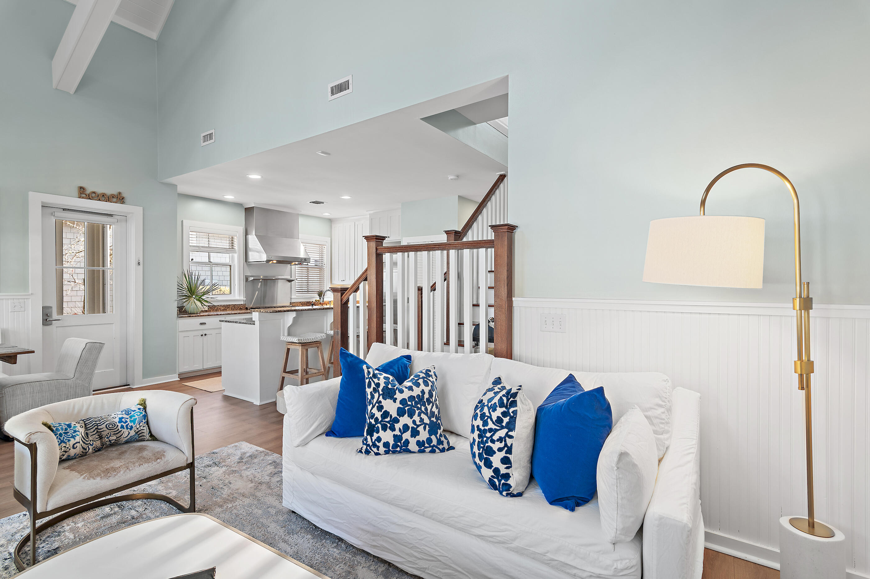 Living room with white sofas and blue pillows, open to a modern kitchen and wood-railed staircase.