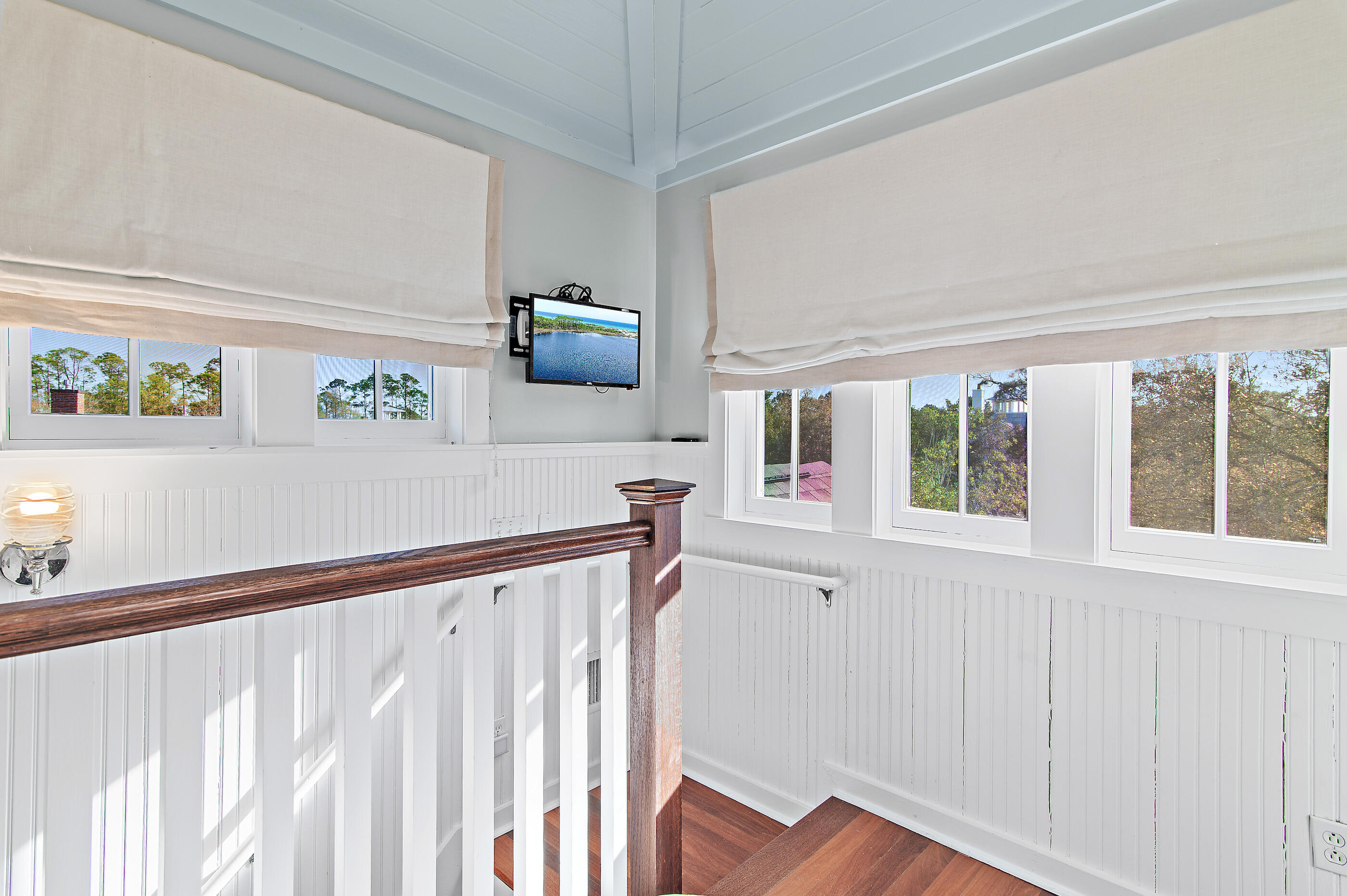 Bright stair landing with white paneled walls, wood floors, and a dark wood railing beside multiple windows.
