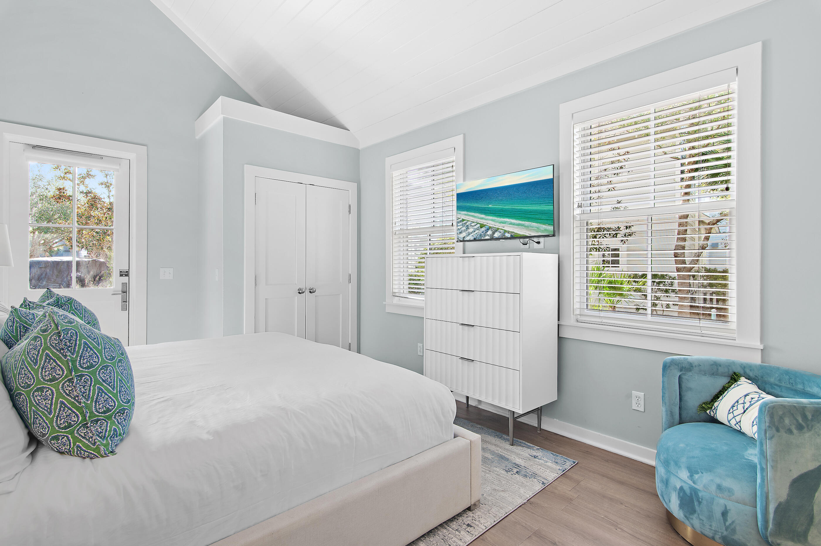 Staged bedroom with a vaulted ceiling, white bed, large windows, and a blue accent chair.