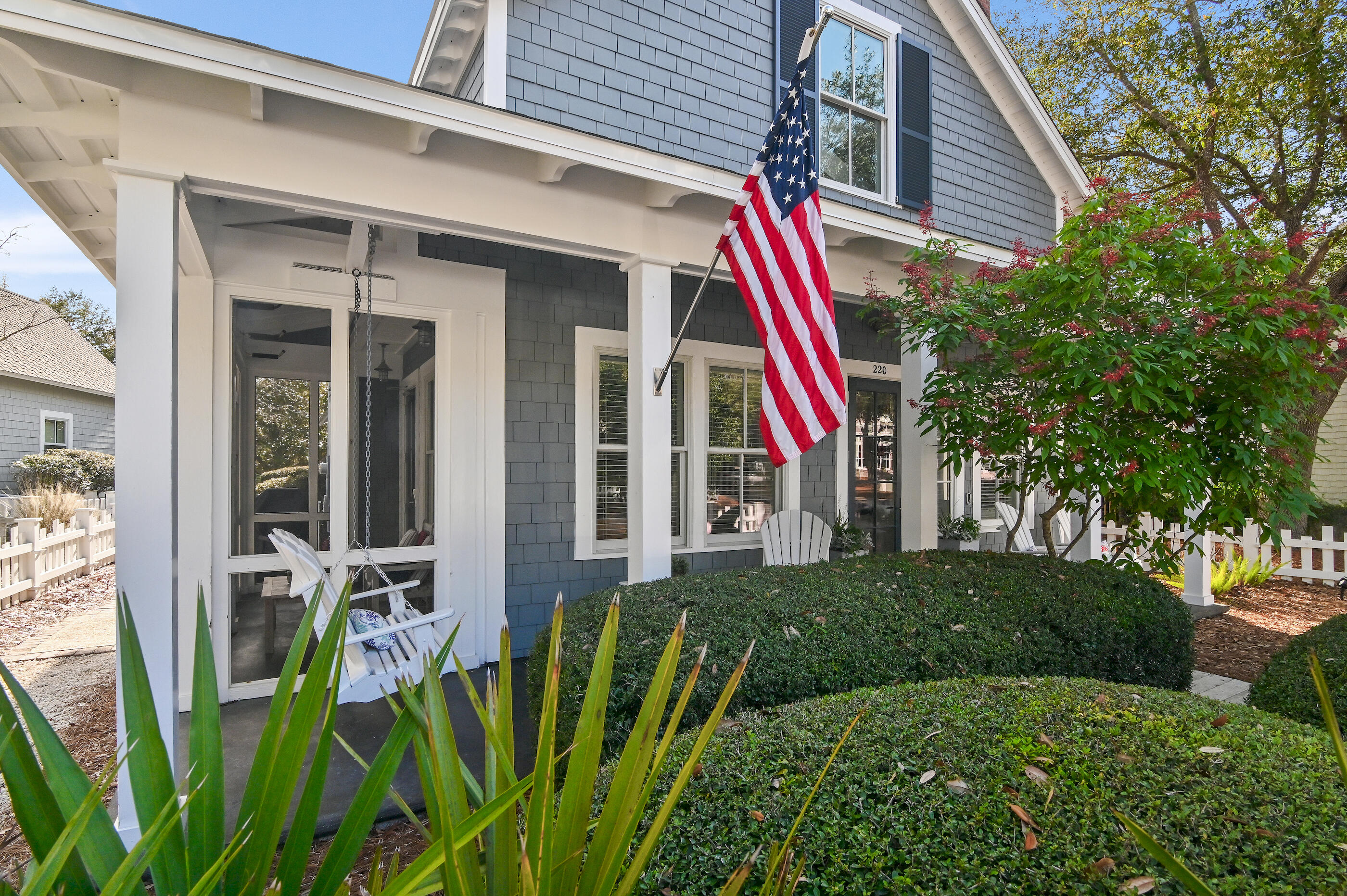 Gray-shingle two-story home with a covered porch, American flag, and landscaped front garden.
