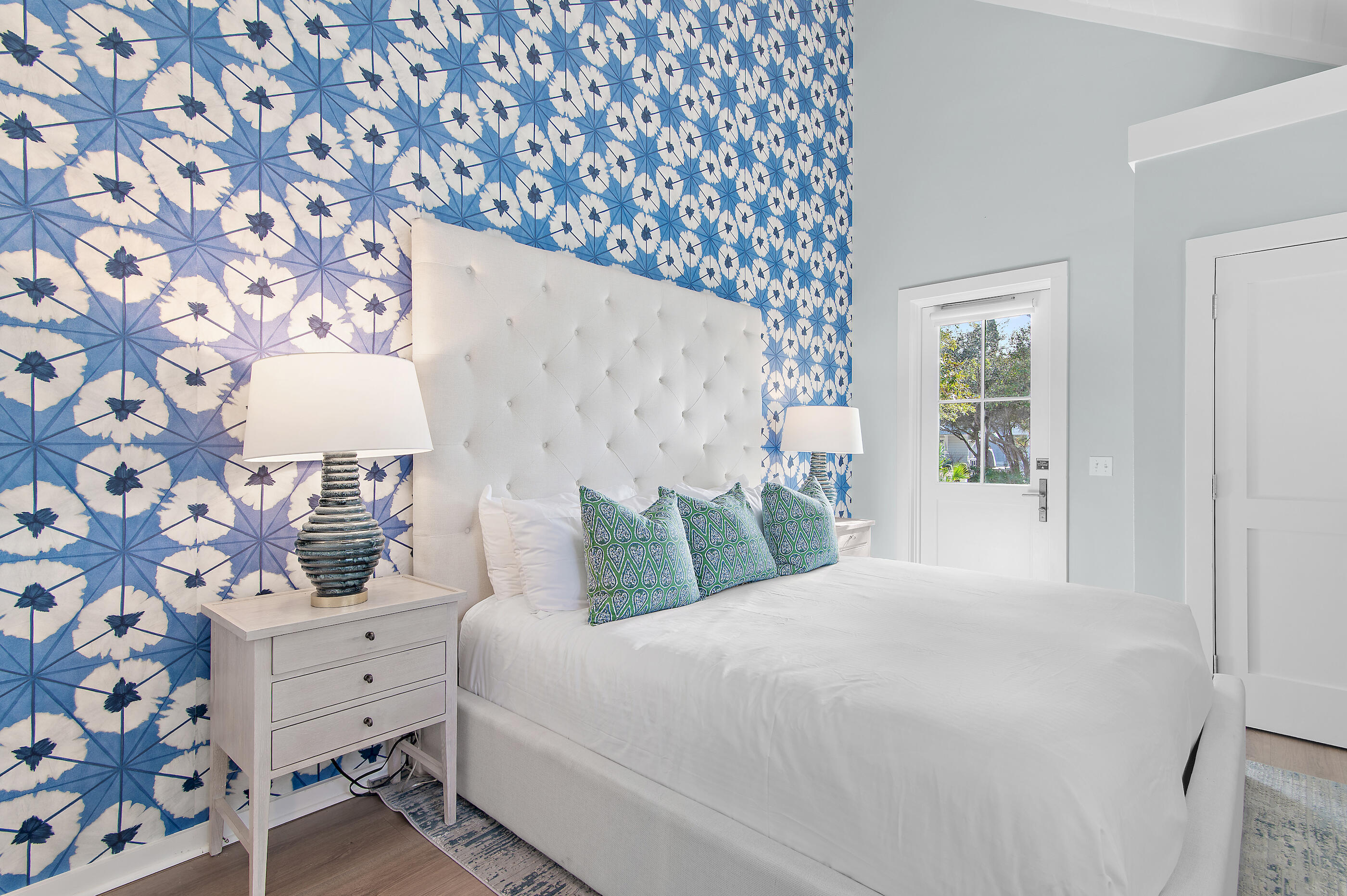 Bedroom with a white tufted bed against bold blue patterned wallpaper and a windowed door.