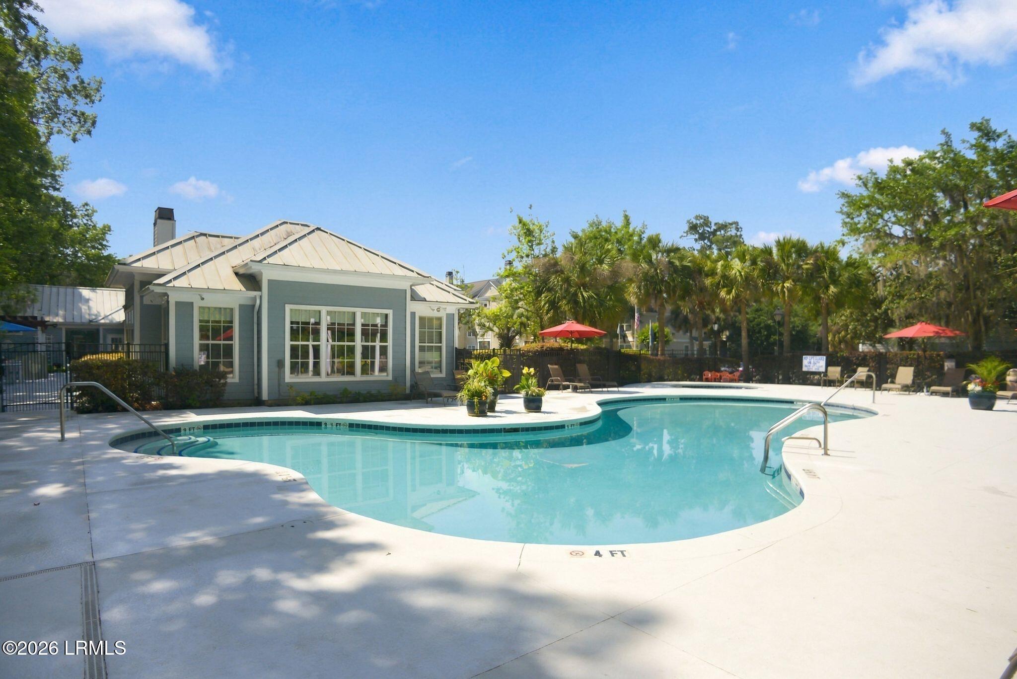 Outdoor pool with a curved shape, lounge chairs, red umbrellas, and palms beside a clubhouse.