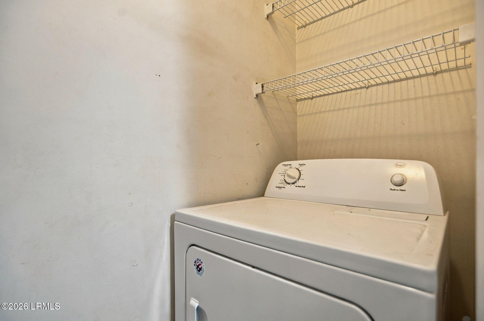 Top-loading washer beneath wire storage shelves in a laundry room.