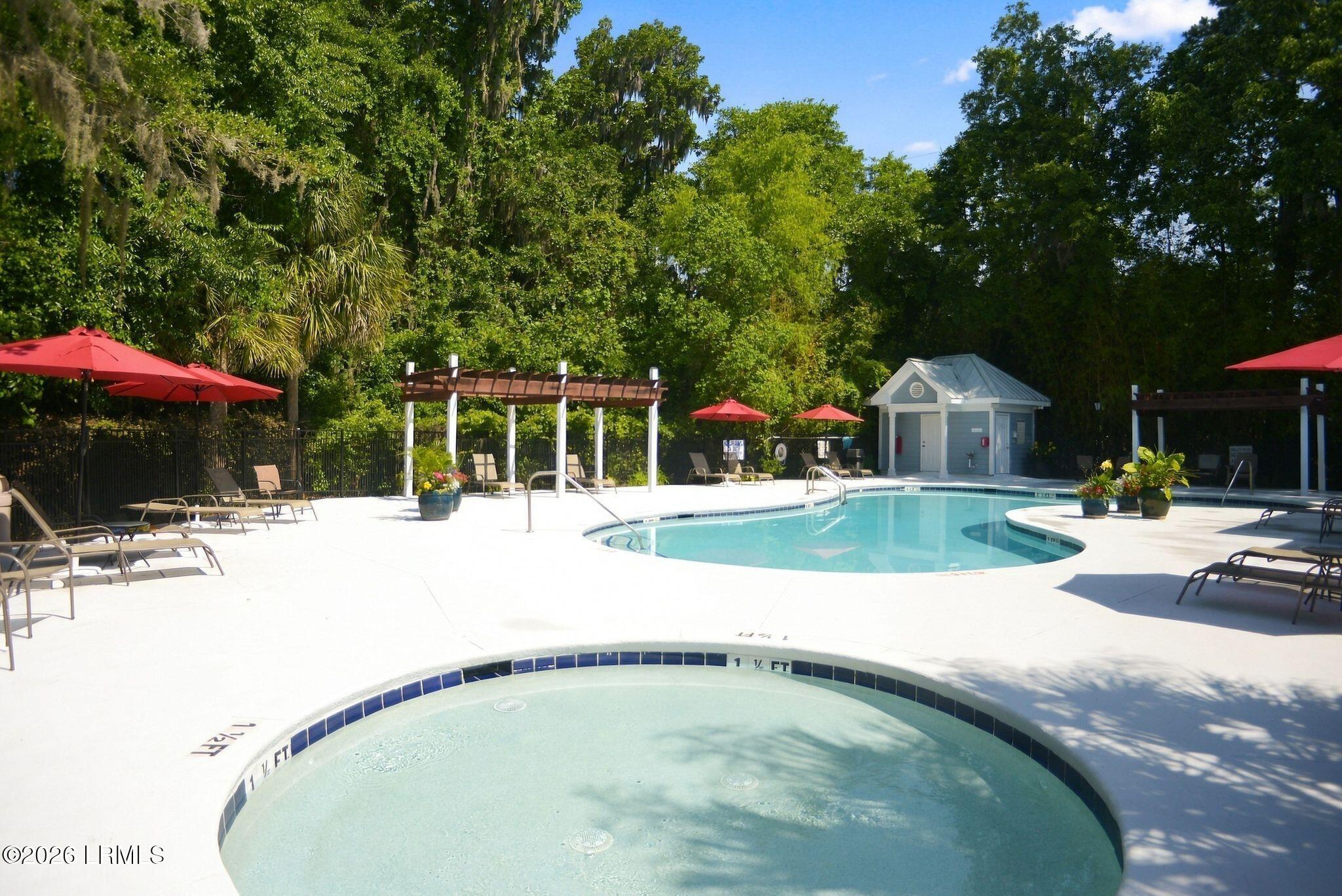 Community pool with a shallow wading area, red umbrellas, and lounge chairs beneath surrounding trees.