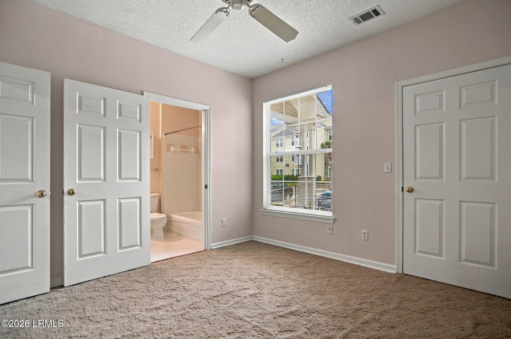 Empty carpeted bedroom with ceiling fan, window, and open doorway to a bathroom.