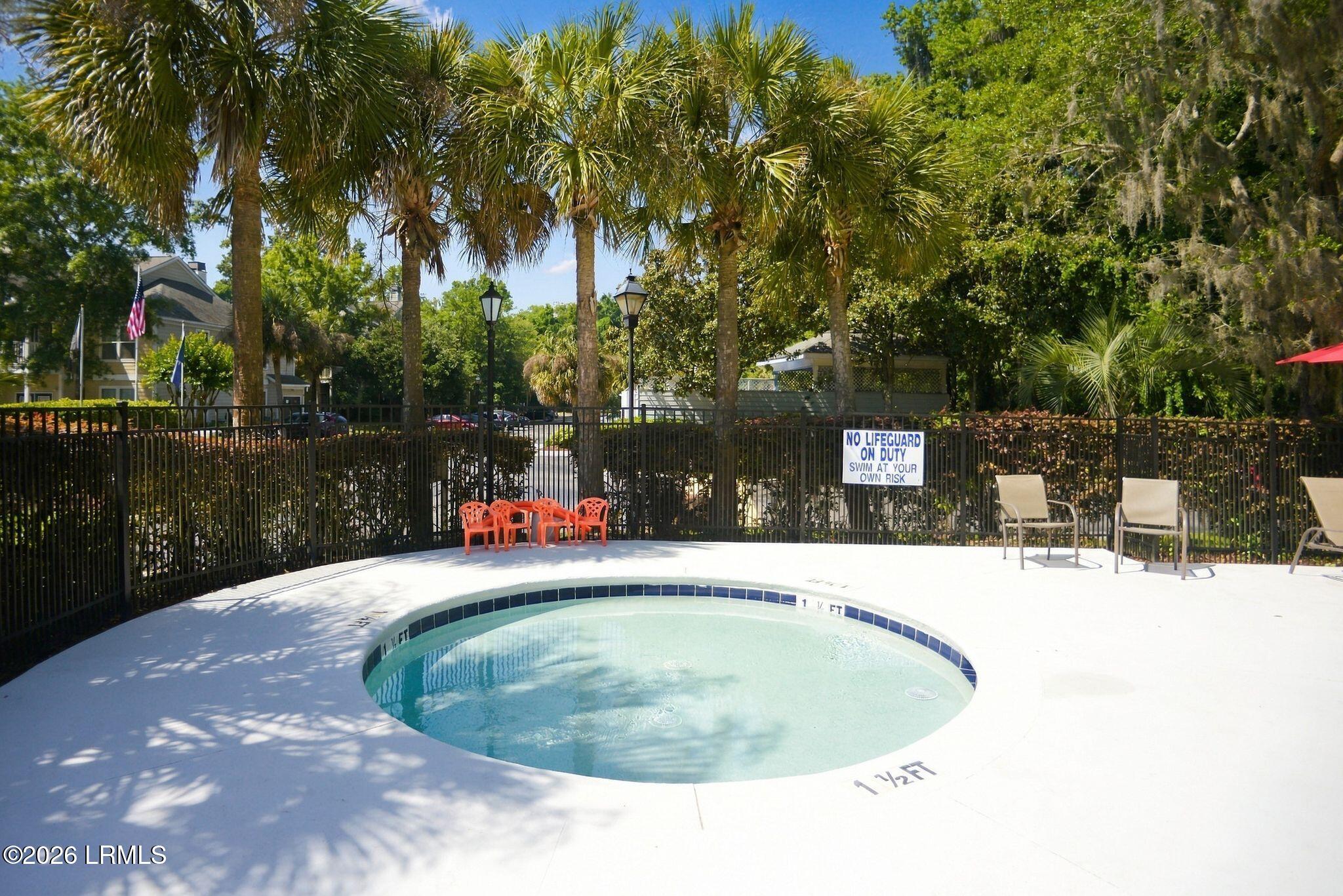 Shallow curved outdoor pool surrounded by palm trees, patio chairs, and a black metal fence.
