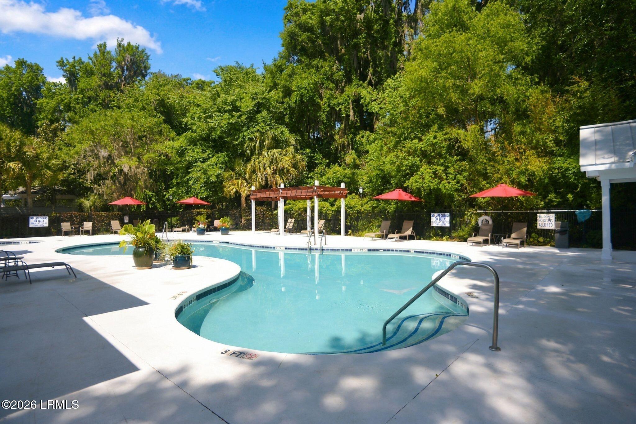 Outdoor pool with a curved shape, red umbrellas, lounge chairs, and a shaded pergola.