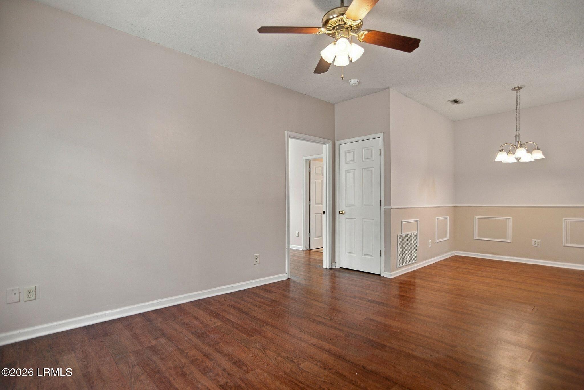Empty dining room with dark wood floors, decorative wall paneling, ceiling fan, and chandelier.