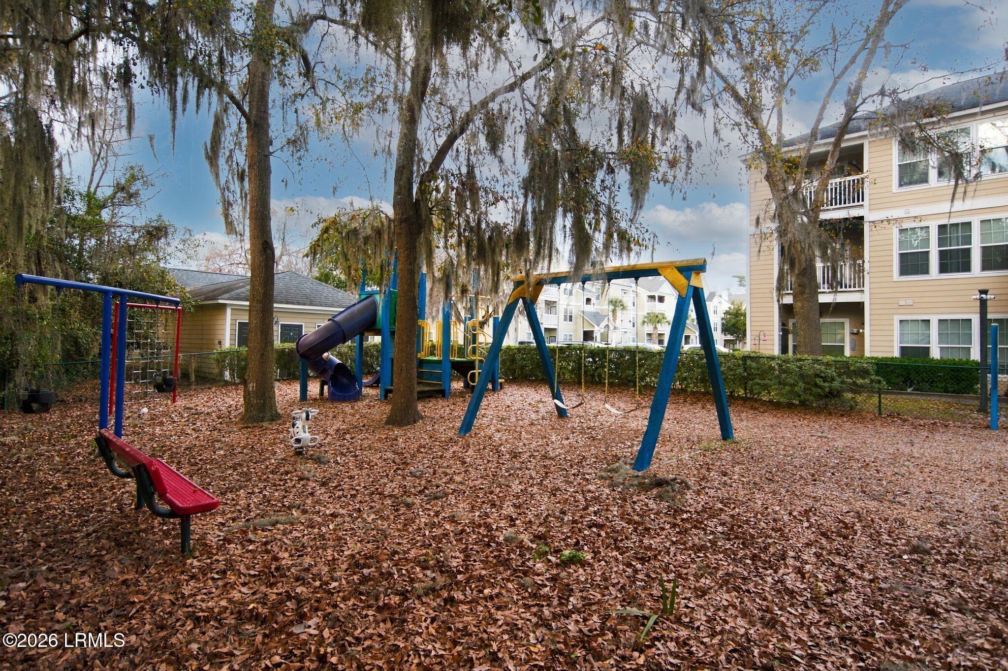 Community playground with swings and a slide beneath moss-draped trees beside apartment buildings.