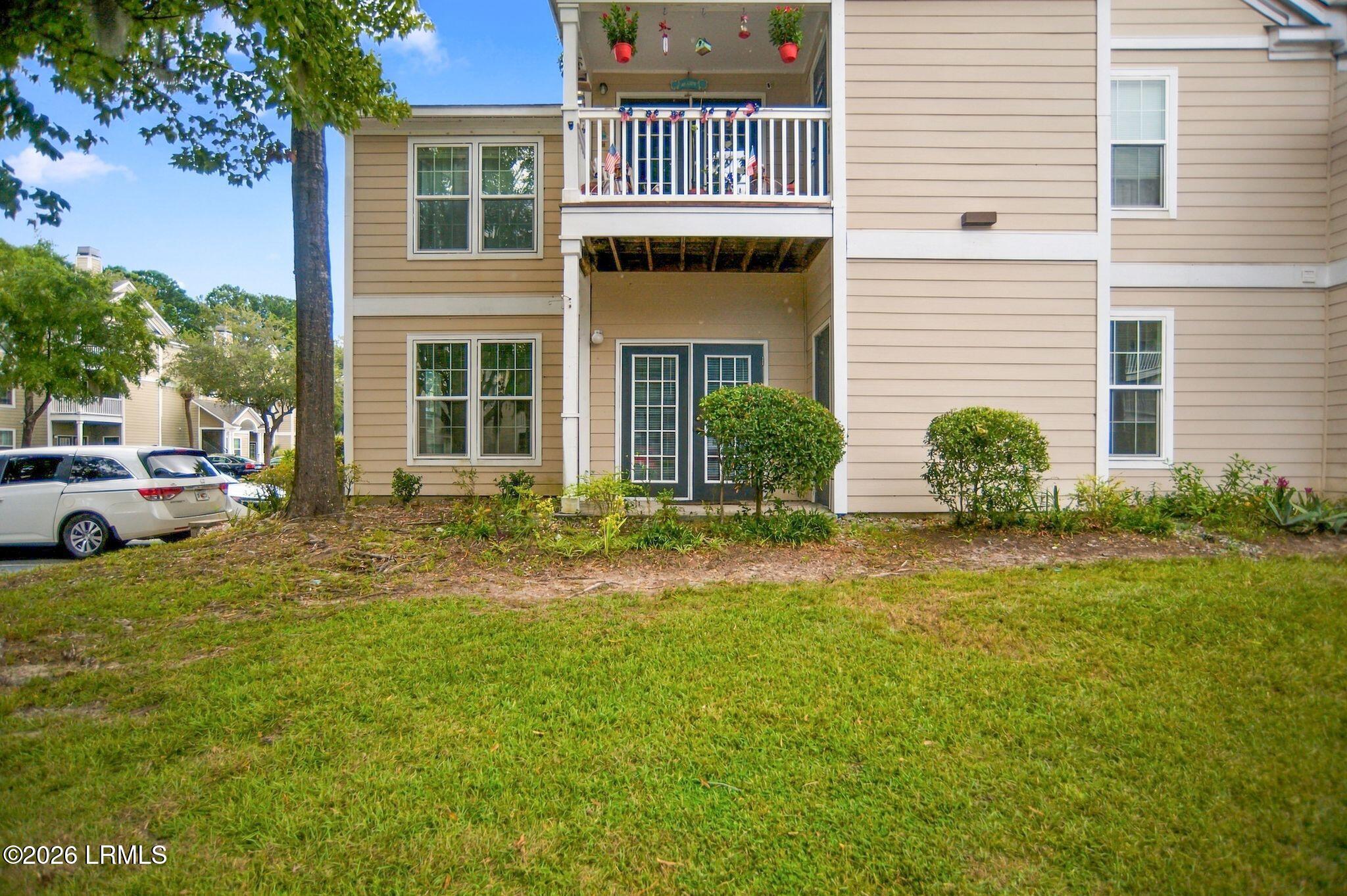 Beige condominium exterior with a covered entry, elevated balcony, shrubs, and lawn.