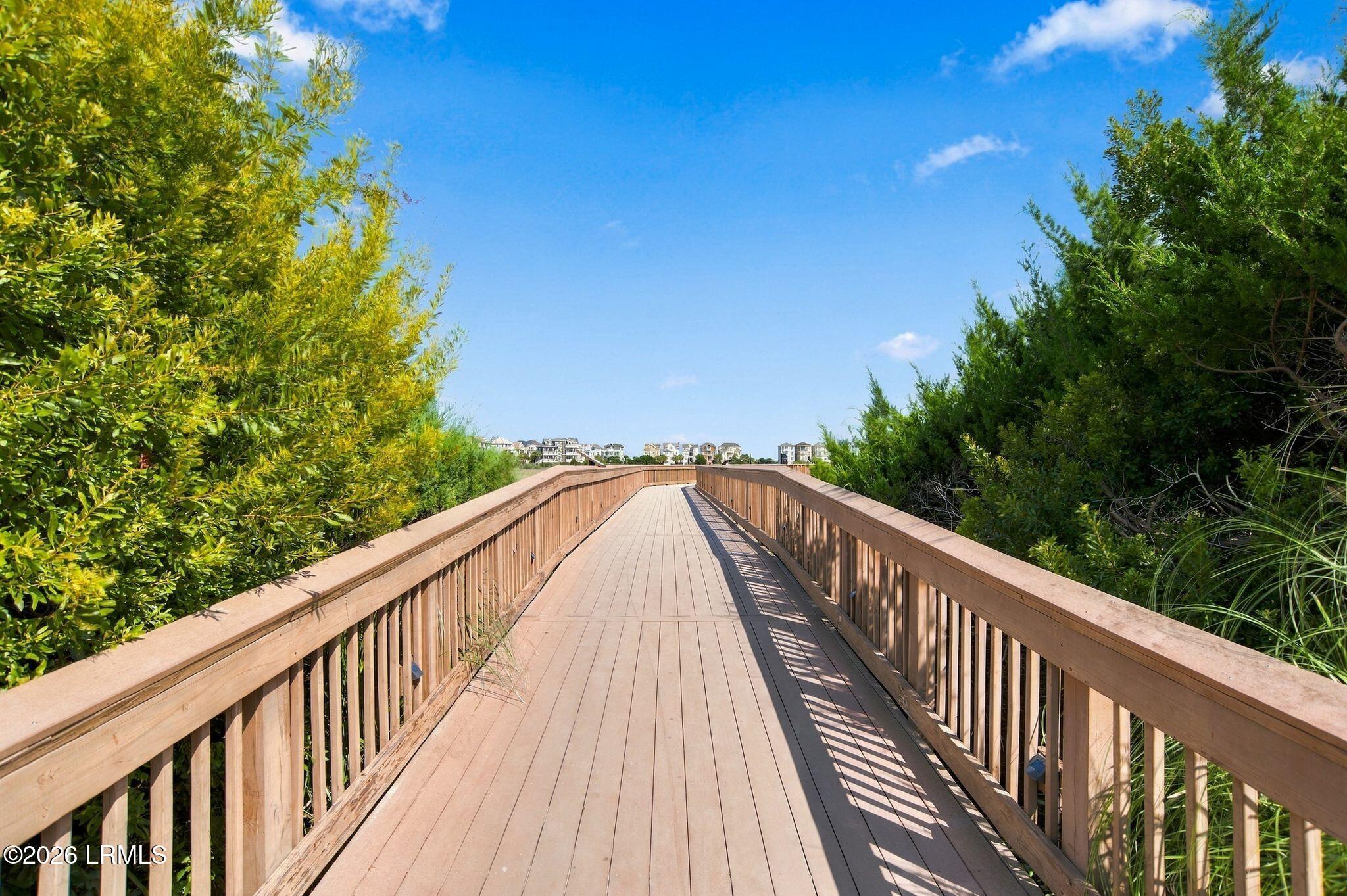 Wooden boardwalk through coastal greenery toward distant buildings.