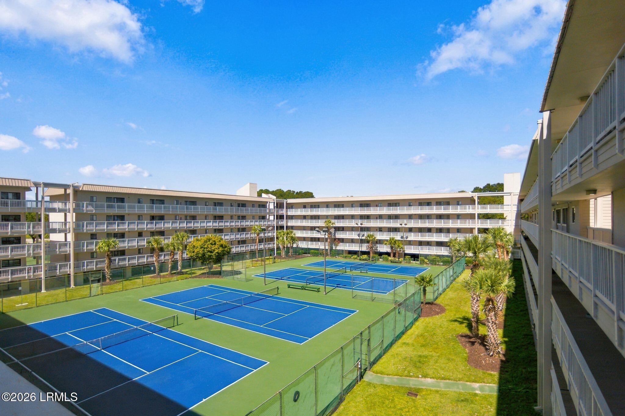 Multiple blue tennis courts in a landscaped courtyard surrounded by condominium buildings and palm trees.