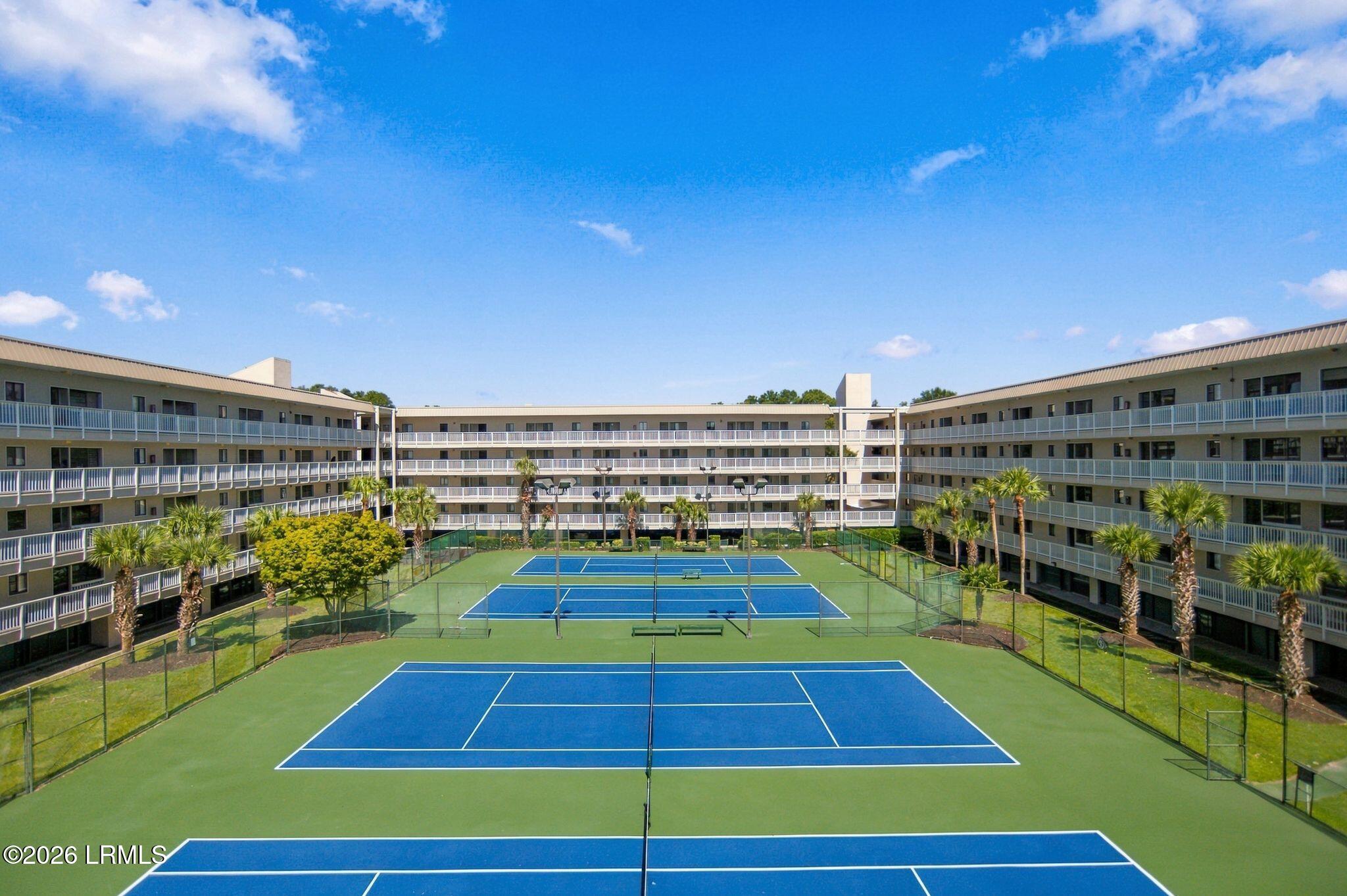 Tennis courts between condominium buildings, with palm trees and white-railed balconies.