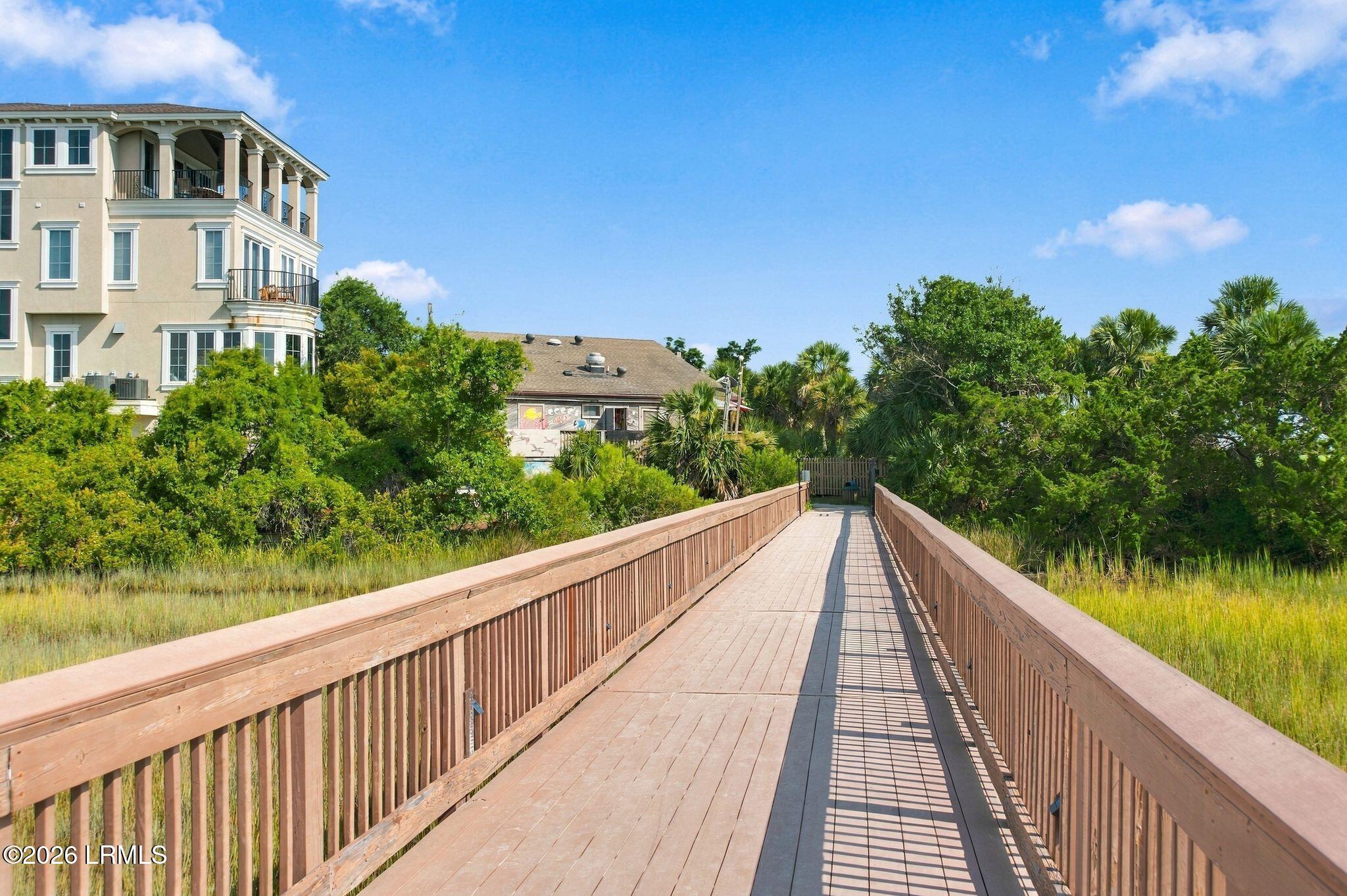 Wooden boardwalk crosses marsh grass toward a gate, with palm trees and a multi-story building nearby.