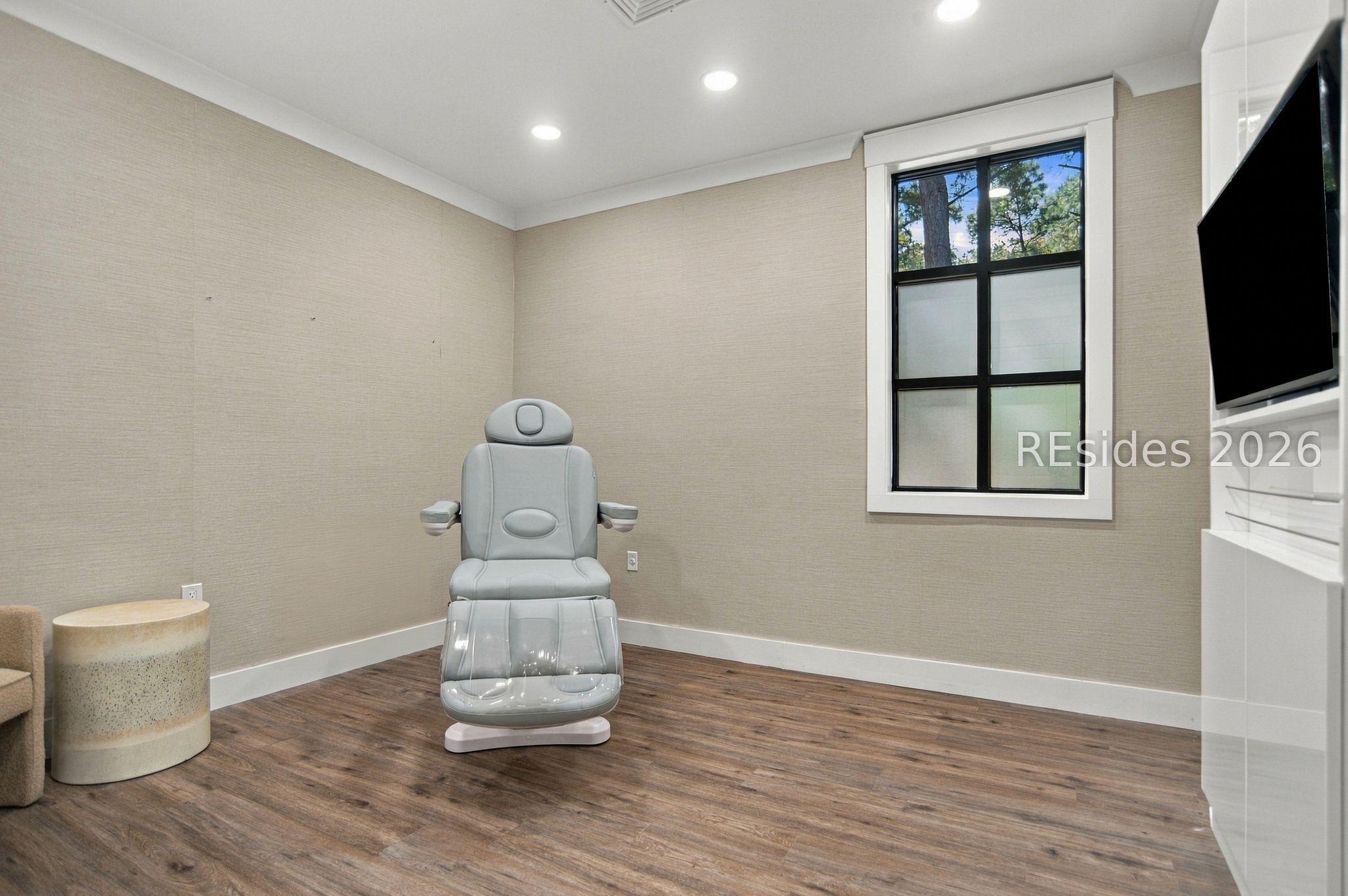 Treatment room with a gray reclining chair, wood-look floors, and a black-framed window.