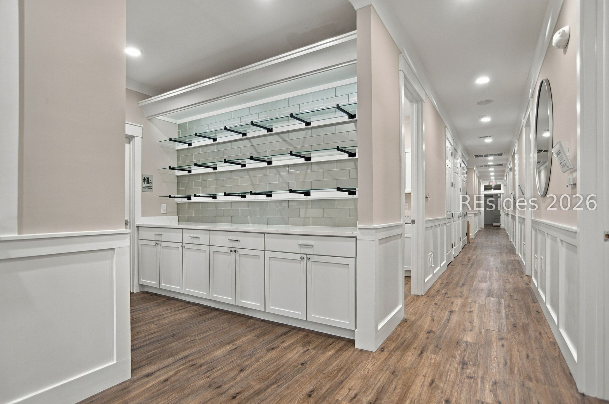 Empty retail interior with glass display shelves, white cabinetry, and a long wood-look-floored hallway.