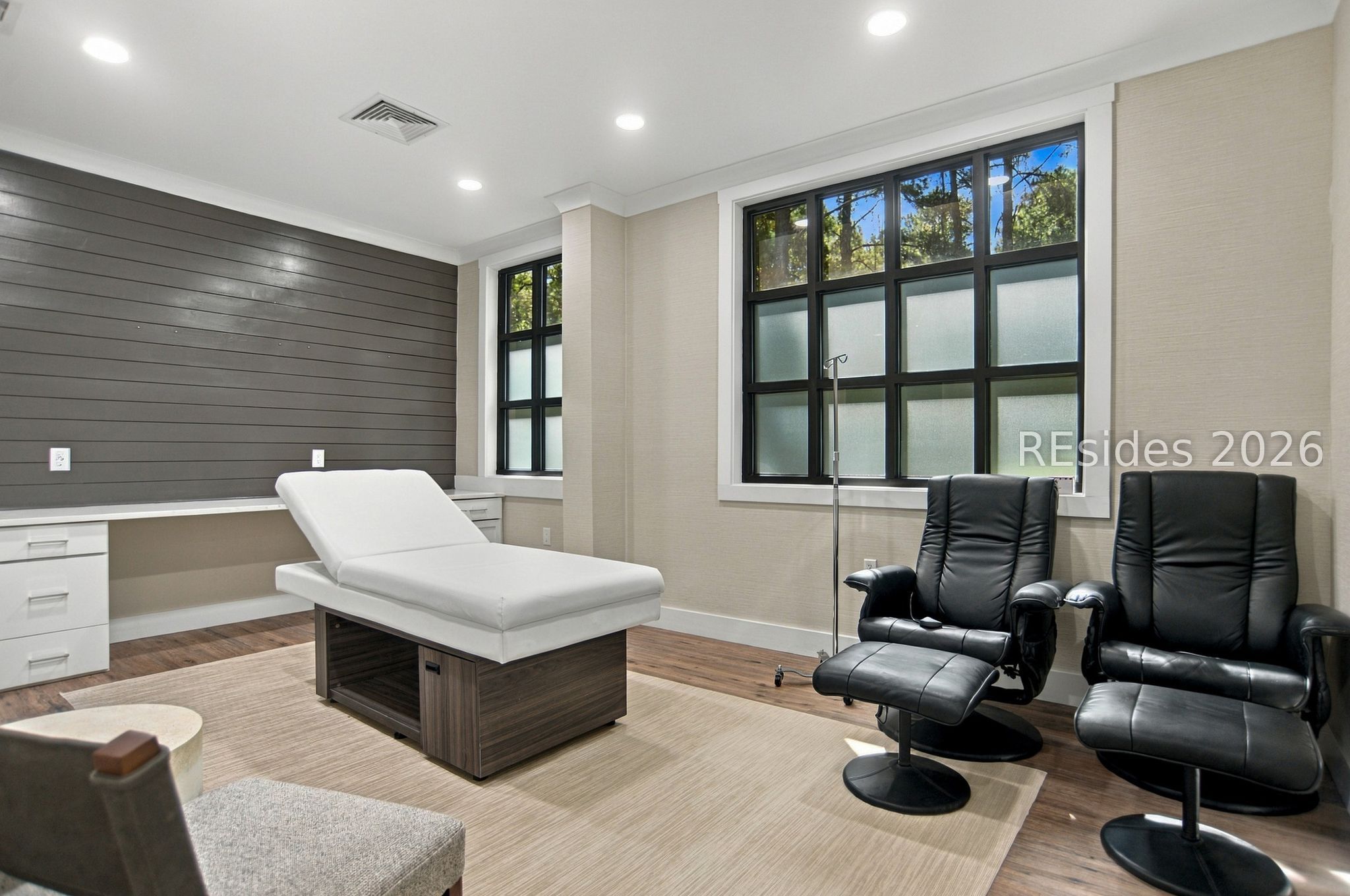 Furnished room with a white treatment table and two black reclining chairs beside large windows.