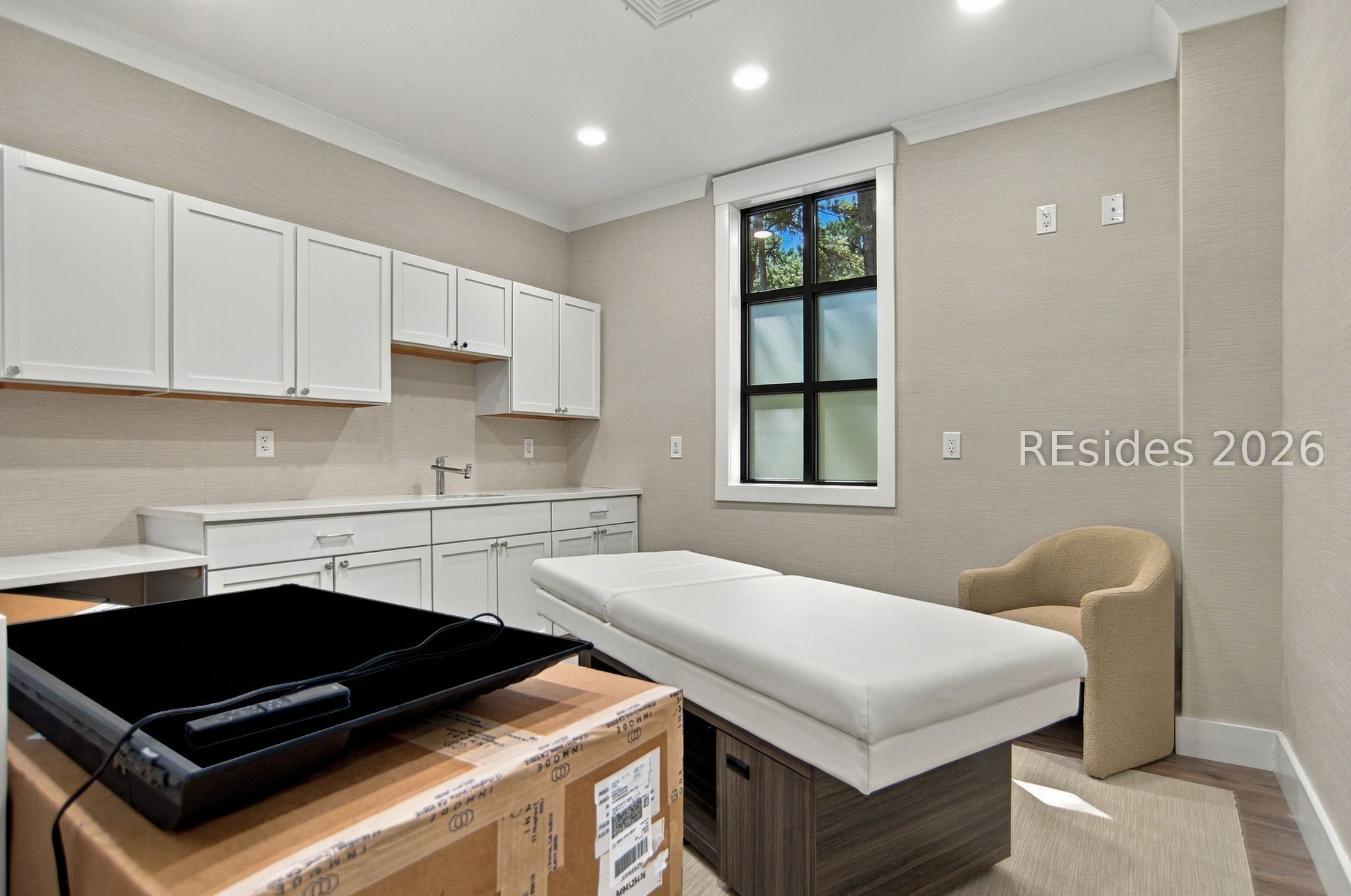 Medical treatment room with a padded examination table, white cabinetry, and a black-framed window.