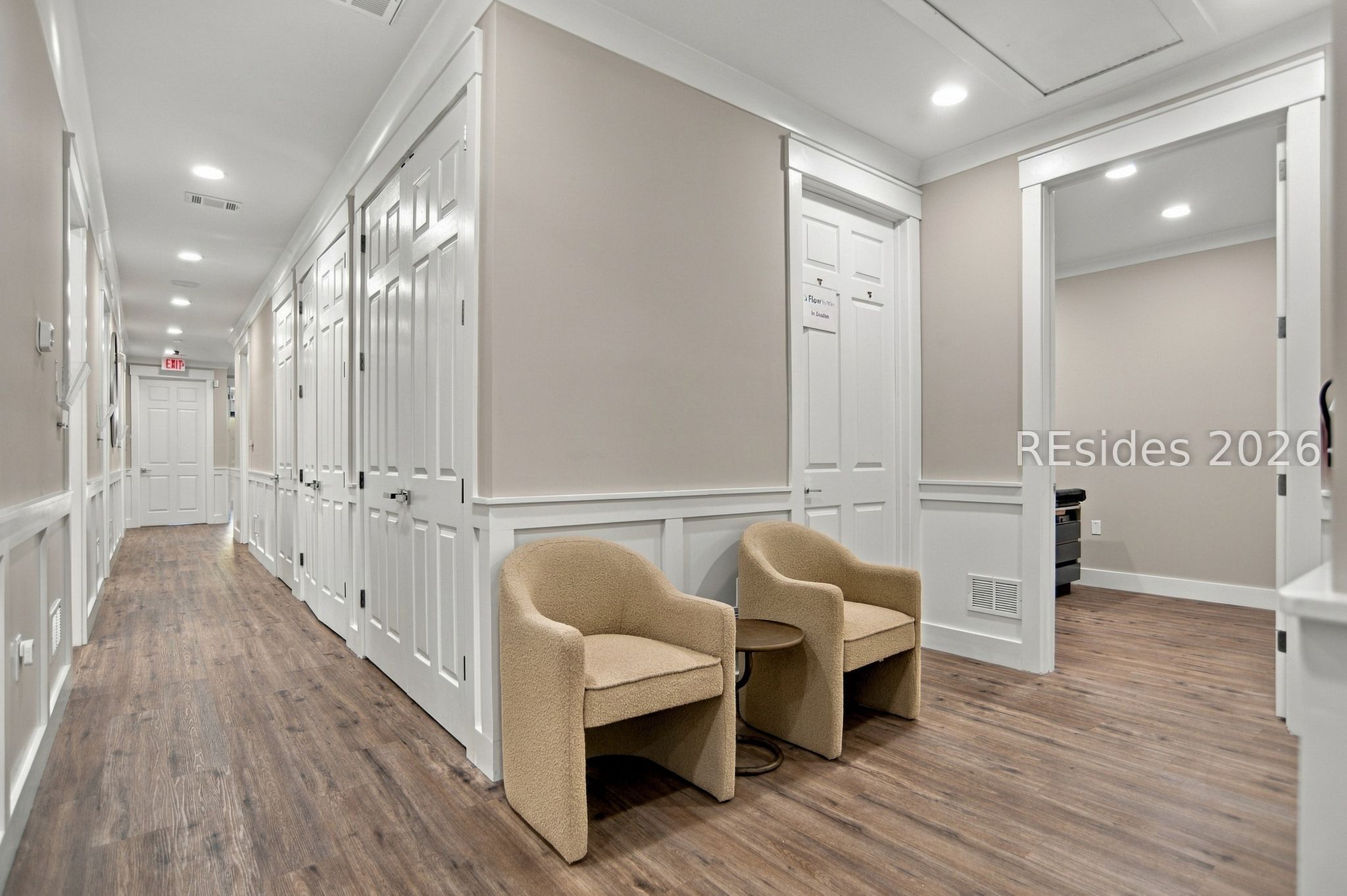 Commercial hallway with white paneled doors, wood-look floors, and two beige chairs beside an open doorway.