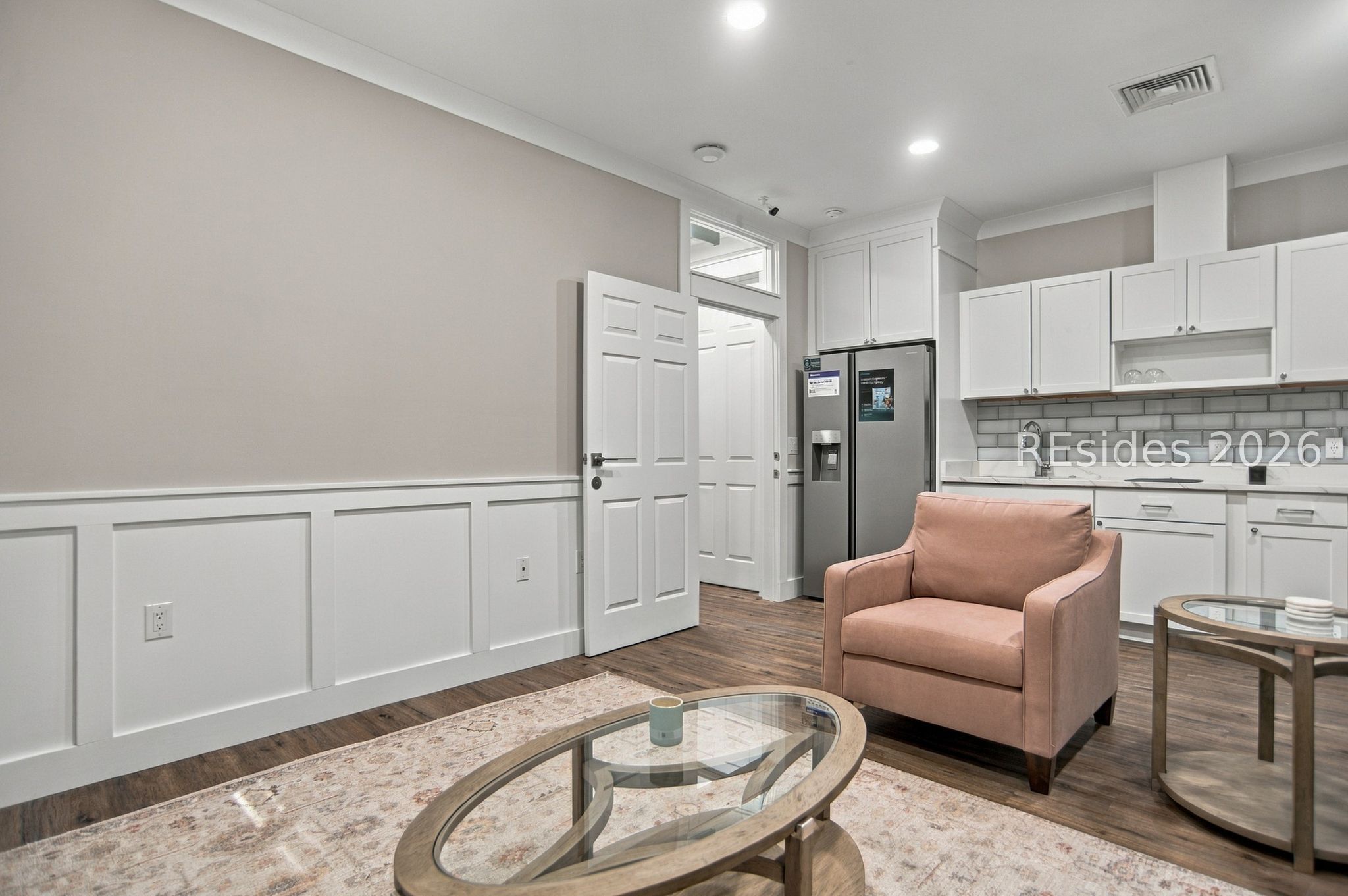 Staged living area with a pink armchair and oval glass coffee table beside a white kitchen.