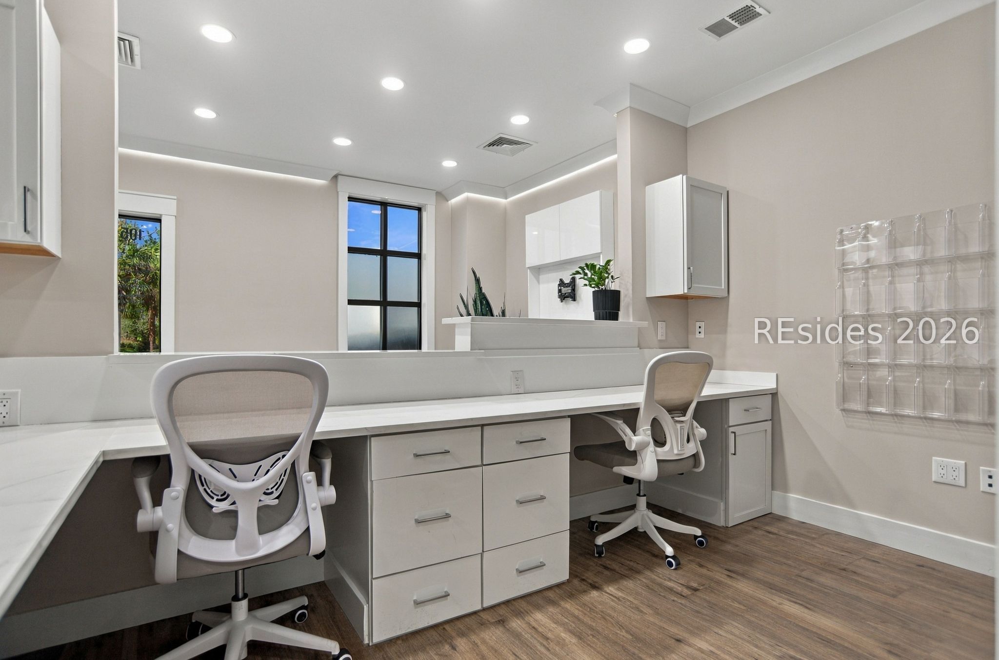 Modern office with a long white workstation, two ergonomic chairs, built-in storage, and large windows.