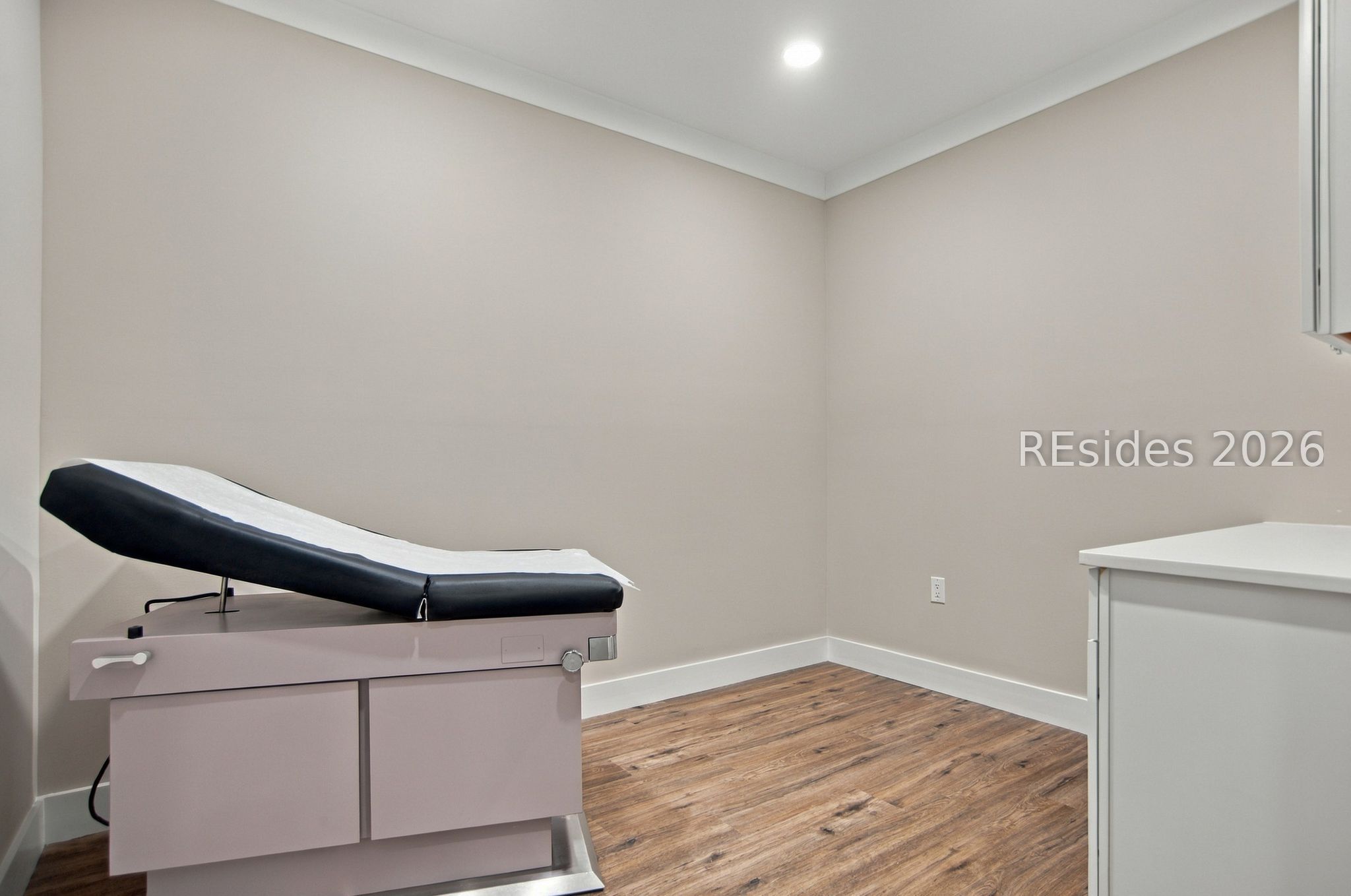 Examination room with an adjustable medical table, white counter, and wood-look flooring.