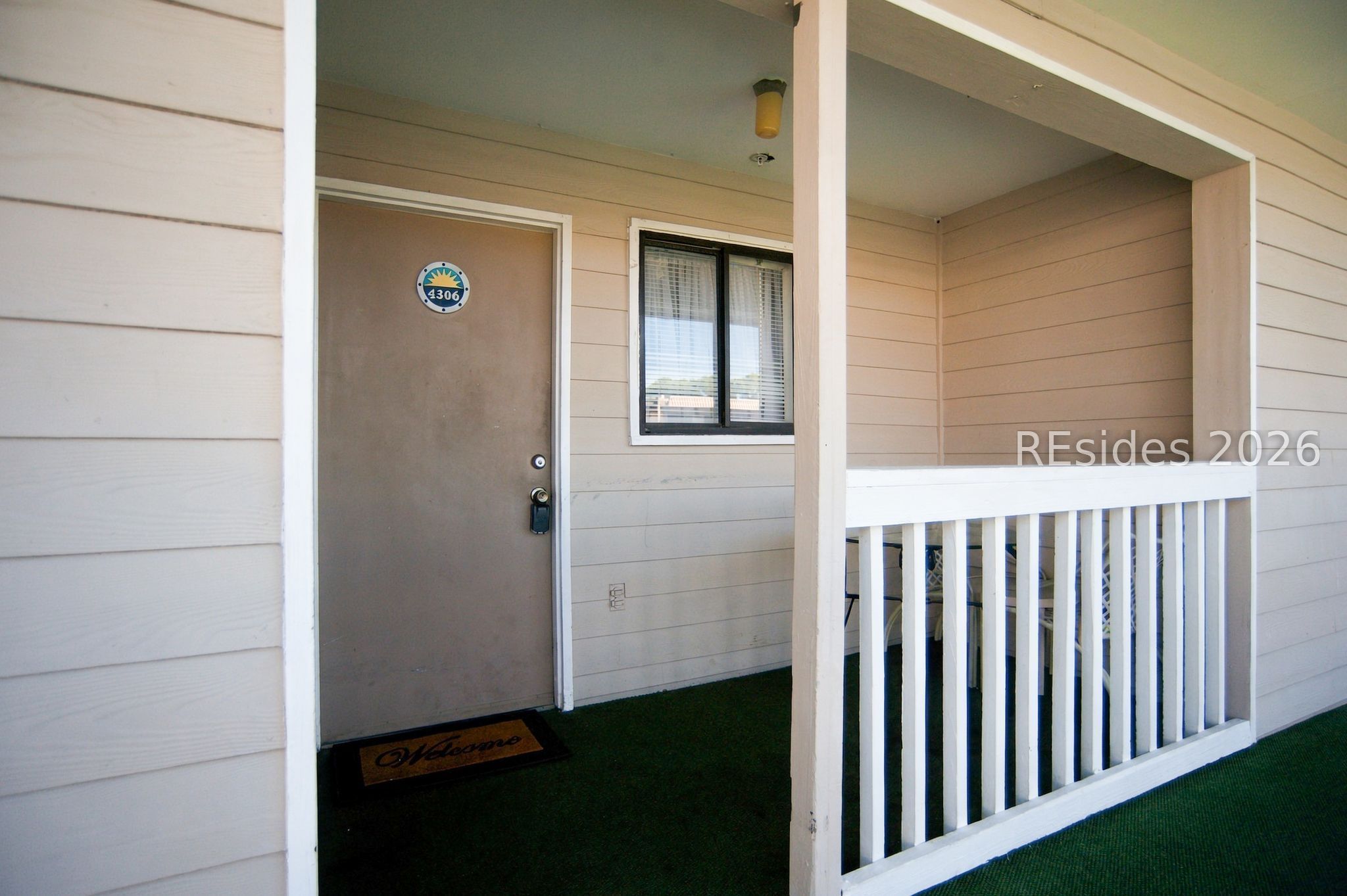 Unit 4306’s covered entrance porch with a brown door, white railing, and green flooring.