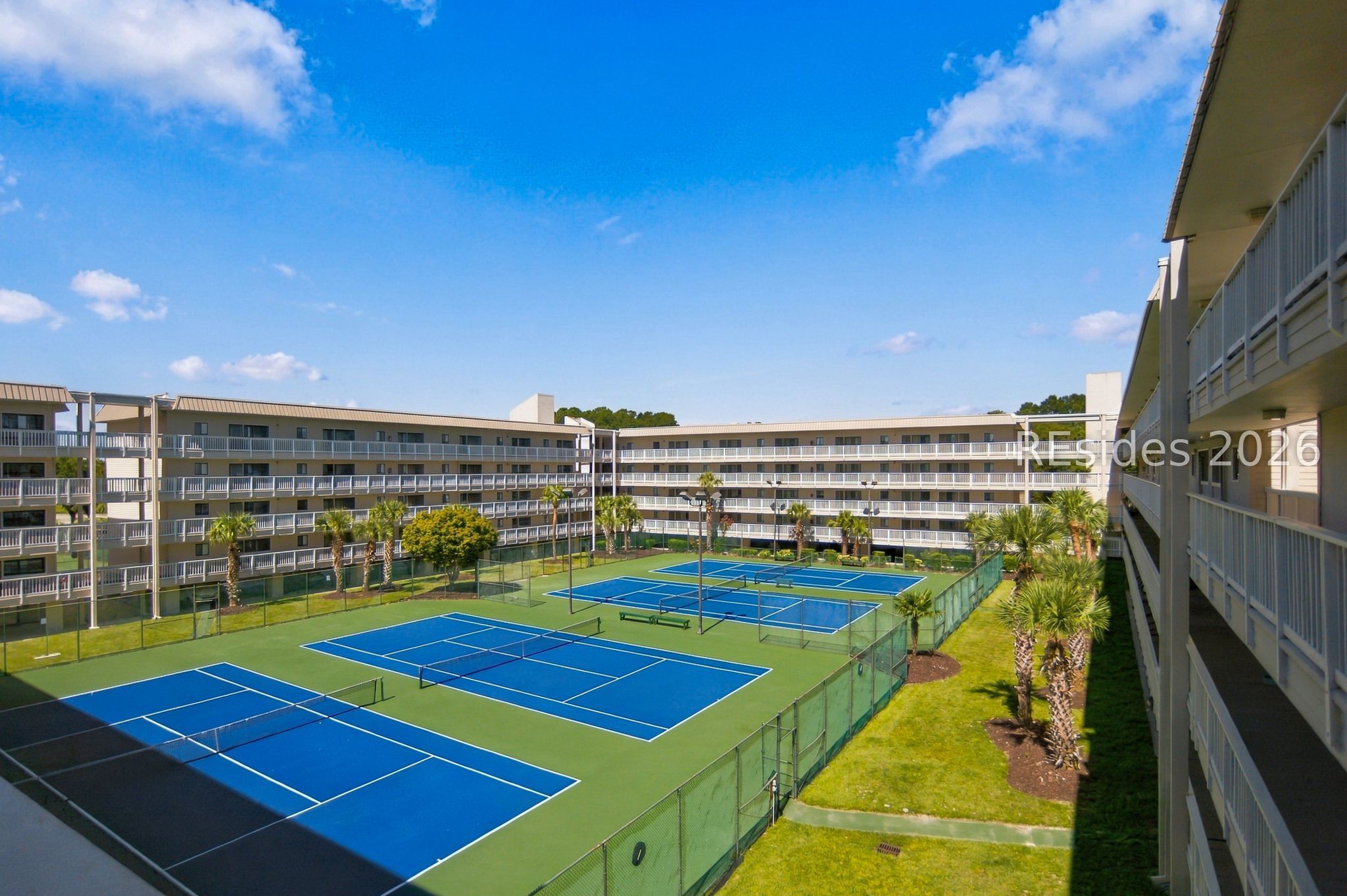 Condominium courtyard with blue tennis courts, palm trees, and balconies.