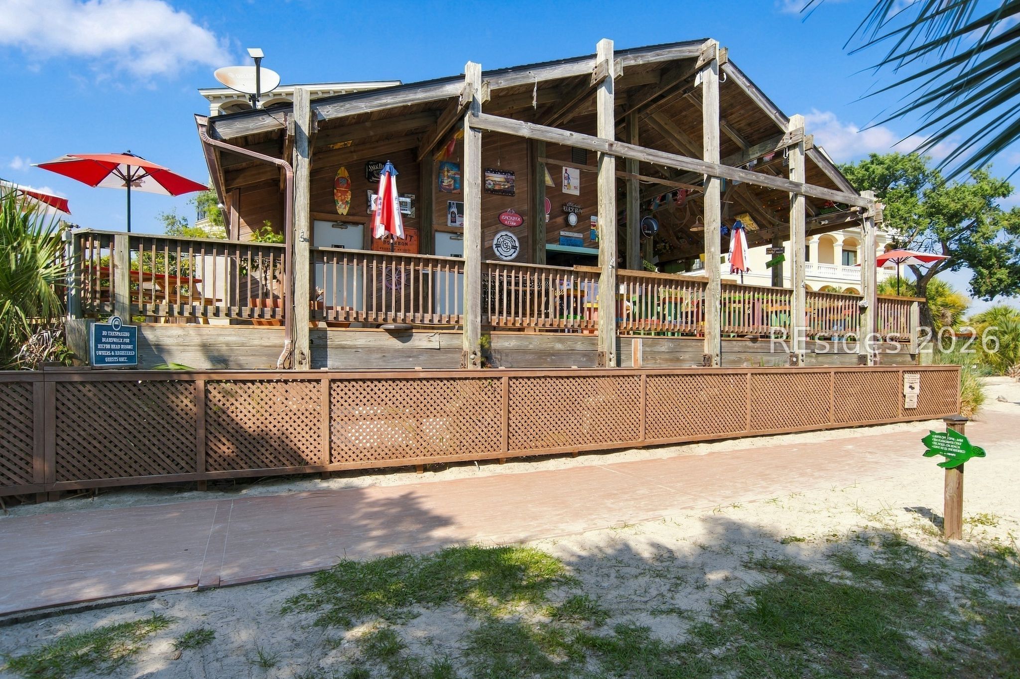 Open-sided wooden pavilion with a raised deck, red umbrellas, and a sandy walkway.