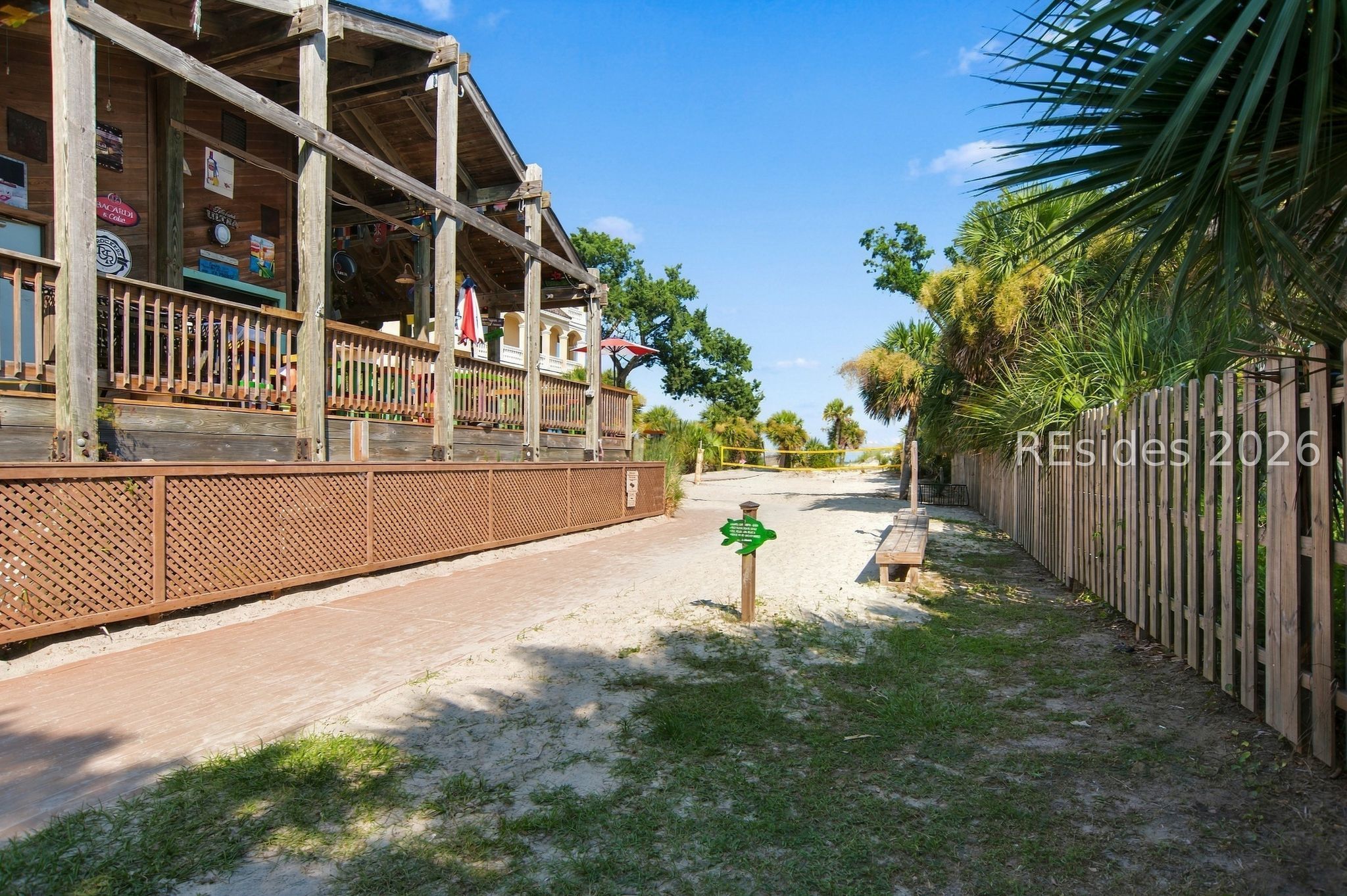 Sandy beach path beside a raised wooden deck, shaded by palms, with water visible in the distance.