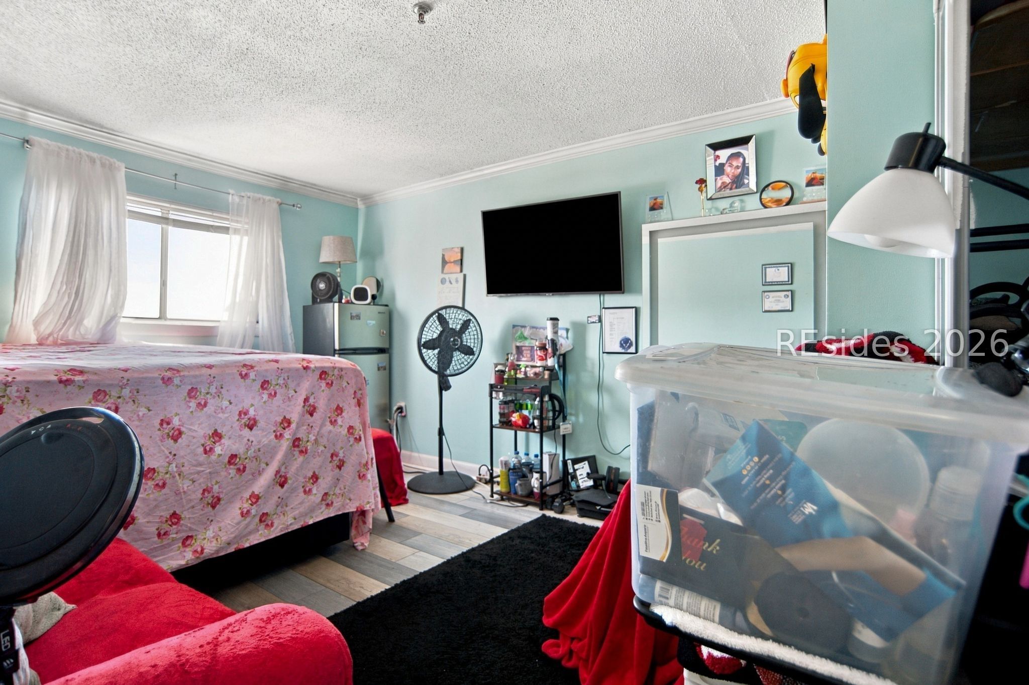 Bedroom with aqua walls, a floral-covered bed, wall-mounted television, and storage bins.