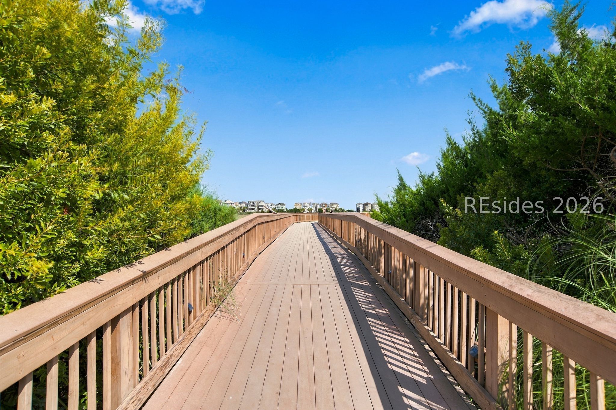 Wooden boardwalk bordered by dense greenery, leading toward buildings in the distance.