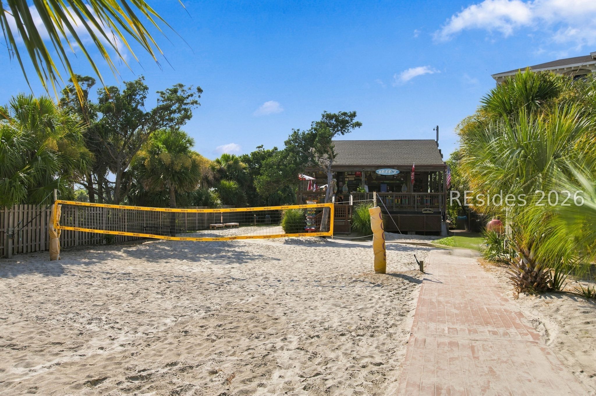 Sandy volleyball court beside a wooden building, framed by palm trees and a brick walkway.