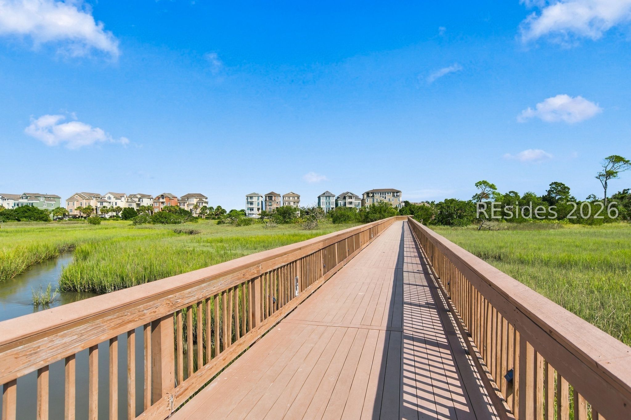 Wooden boardwalk crossing a marsh toward homes, with a narrow water channel alongside.