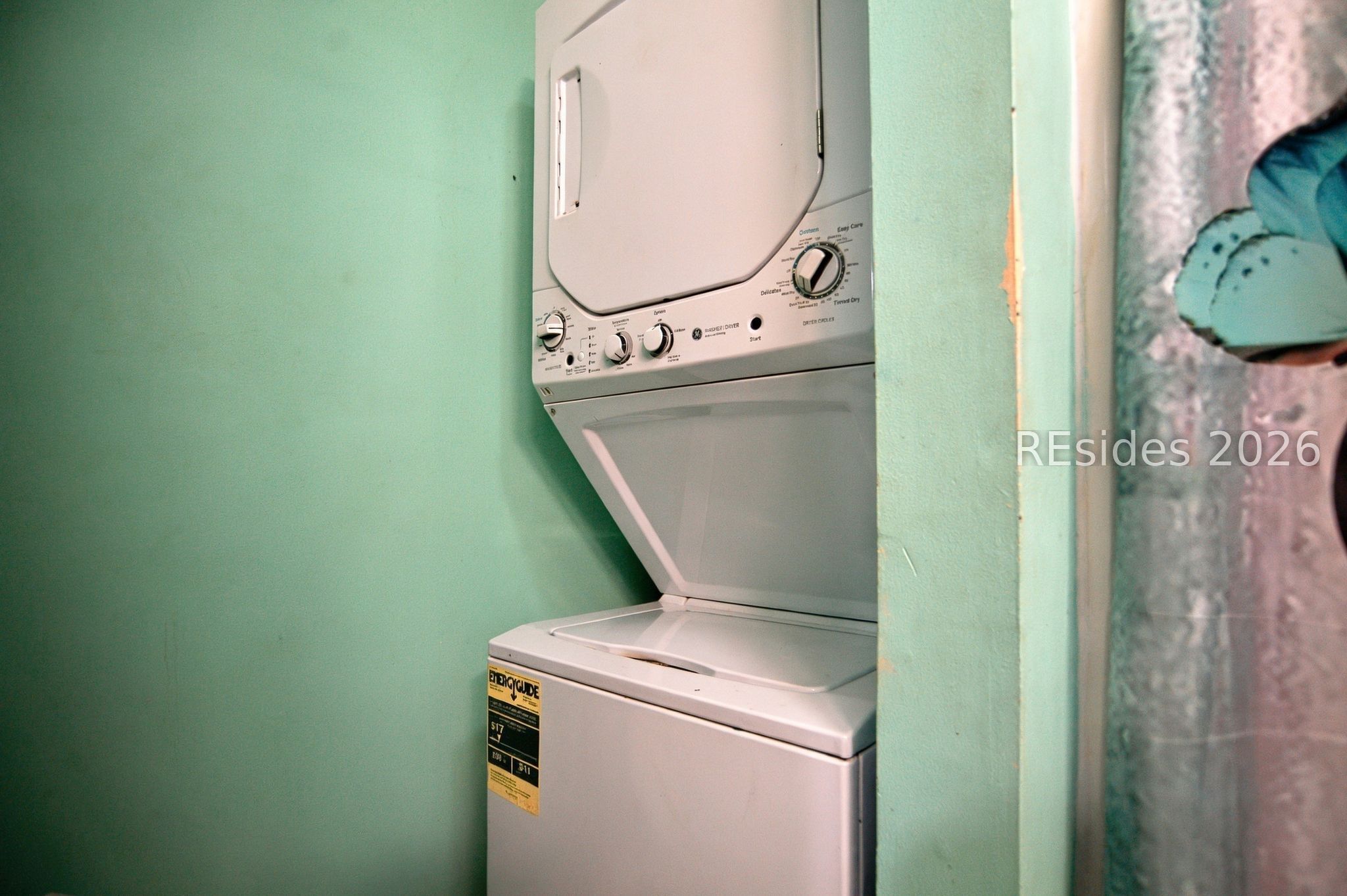Stacked washer and dryer in a narrow mint-green laundry closet beside a patterned curtain.