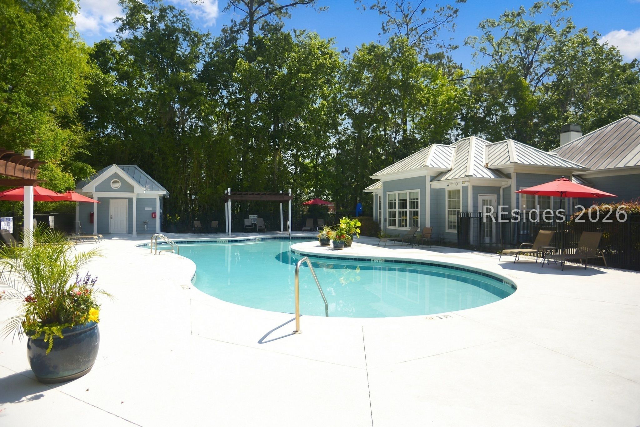 Community pool with a curved turquoise swimming pool, red umbrellas, and a clubhouse beside wooded grounds.